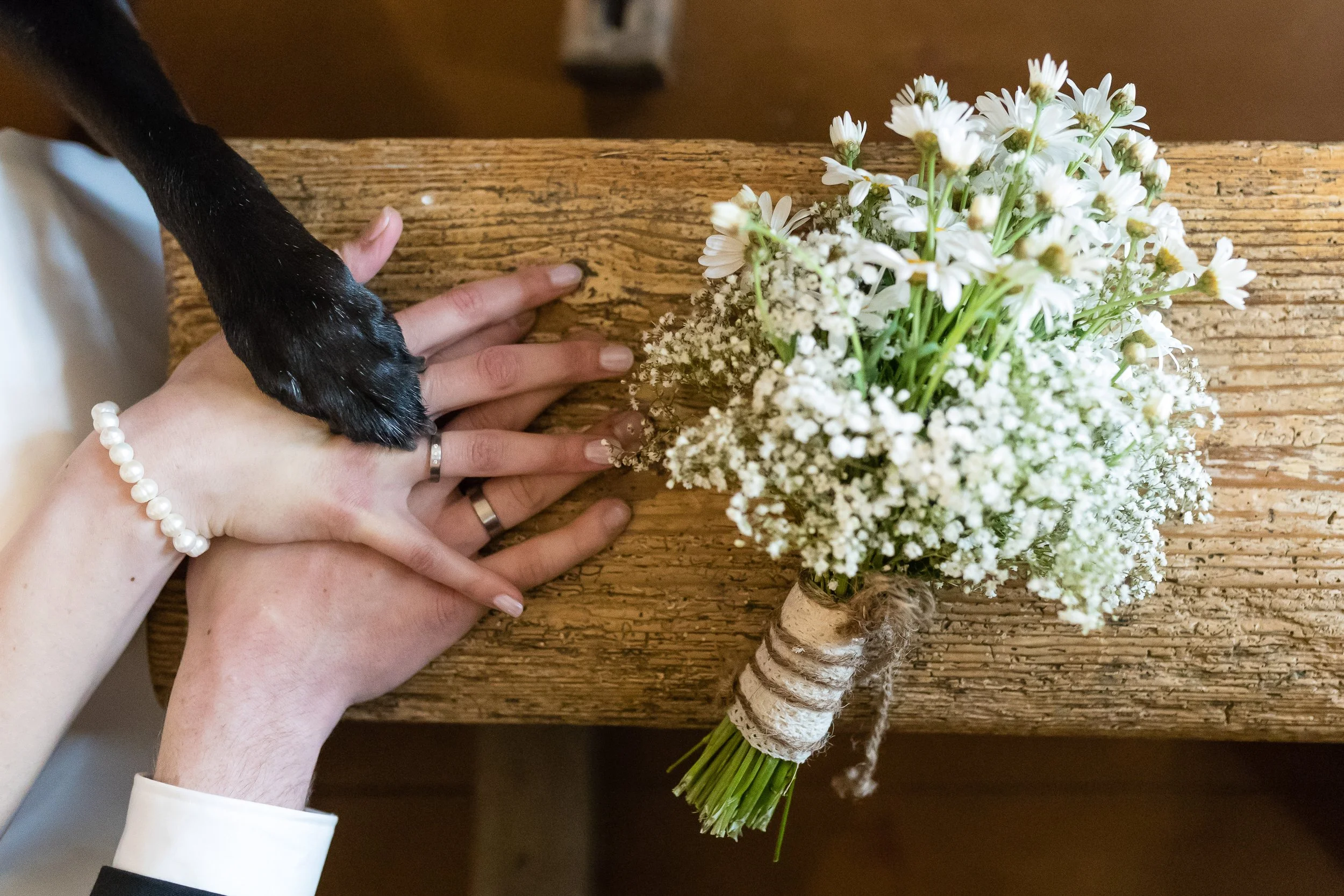 Zwei Hände, eine mit Perlarmband und Ehering, die auf einen rustikalen Holzschreibtisch legen, neben einem Bündel weißer Blumen mit Waldorf-Optik auf dem Tisch.