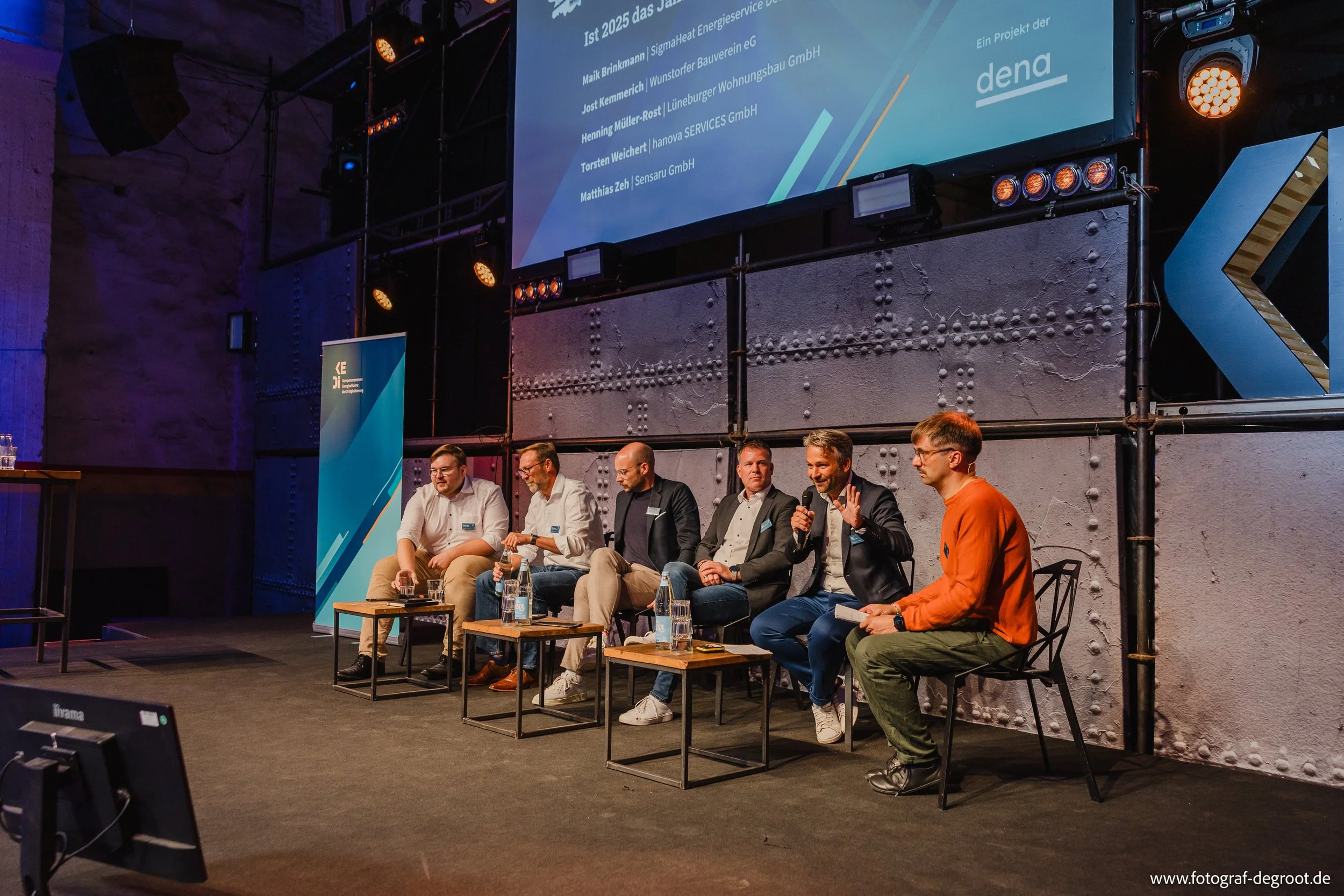 Vier Männer und eine Frau sitzen auf einer Bühne bei einer Podiumsdiskussion bei einer Konferenz, mit großem Bildschirm im Hintergrund, der eine Präsentation zeigt.