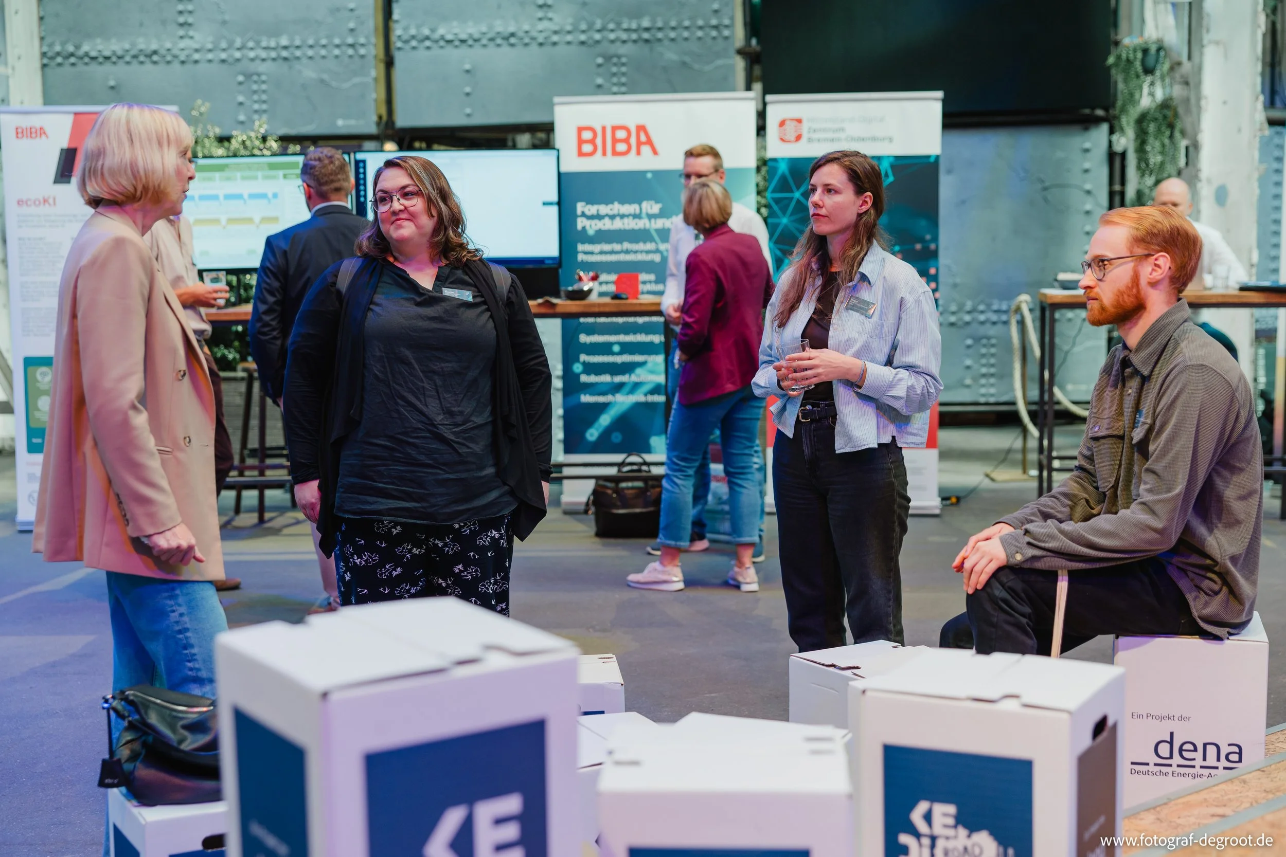 Menschen im Gespräch auf einer Messe mit Informationsständen und Bildschirmen im Hintergrund.