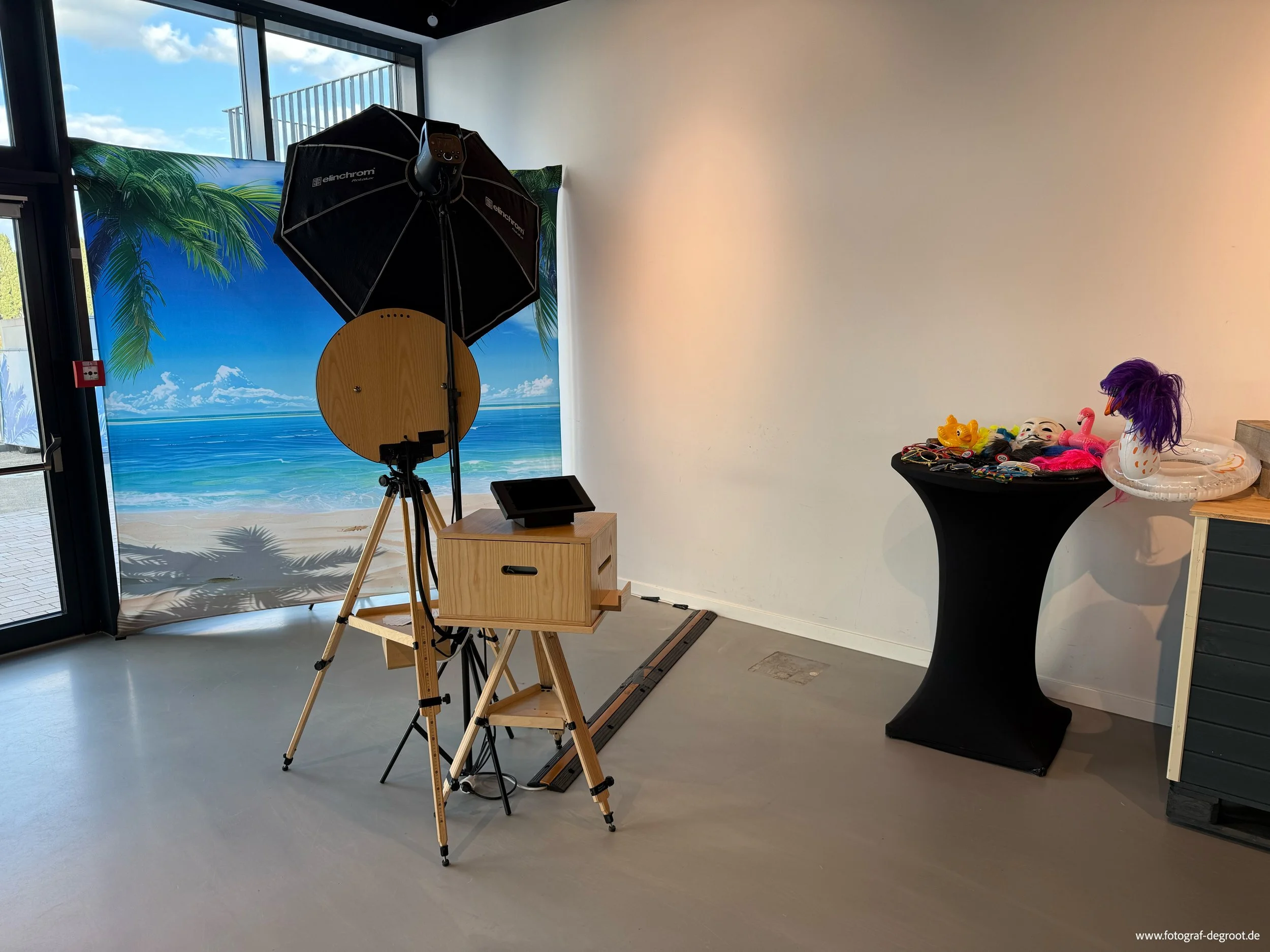 Fotostudio mit Background im Strand-Design, Beleuchtungs- und Kameratechnik, sowie Dekorationen wie eine Flamingo- und eine Hai-Attrappe auf einem schwarz bedeckten Tisch.