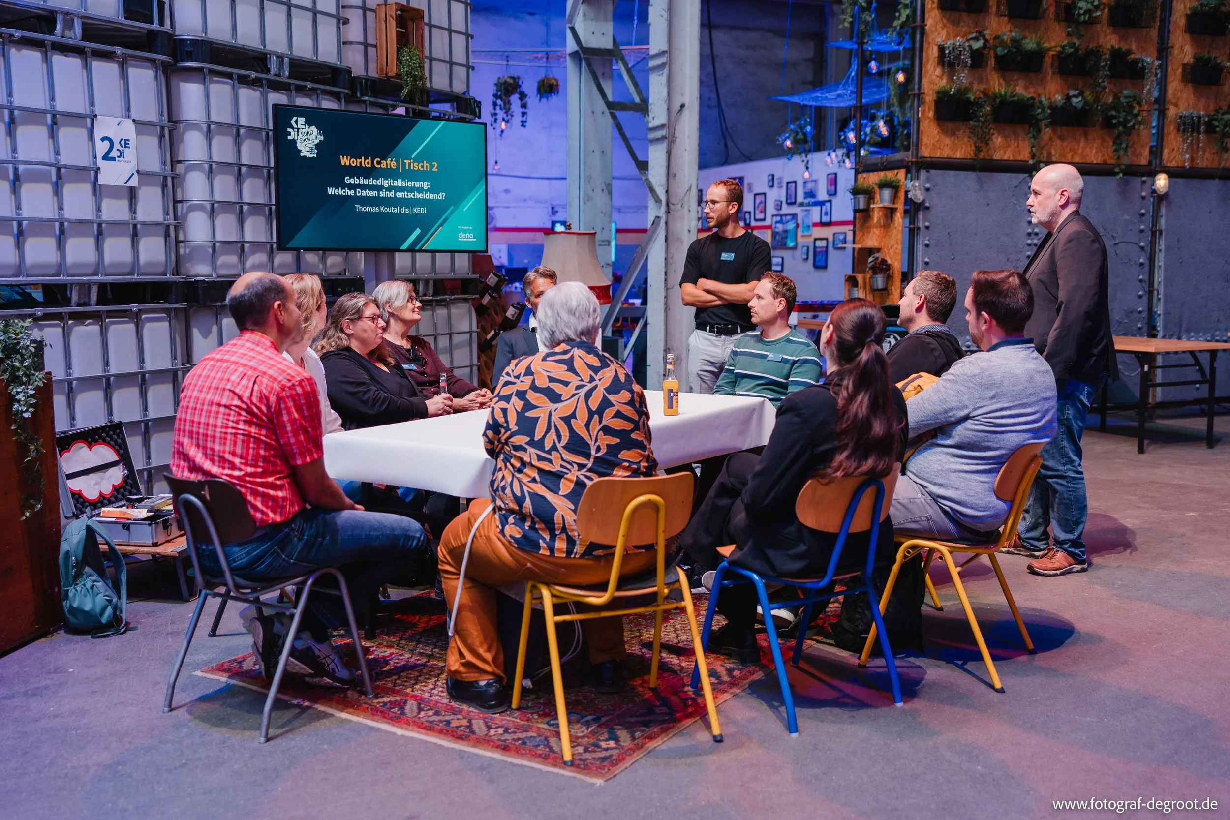 Eine Gruppe von Menschen sitzt um einen langen weißen Tisch in einem modernen Veranstaltungsraum, während ein Mann vor ihnen steht und spricht. Es gibt einen großen Bildschirm an der Wand, auf dem „World Café | Tisch 2“ und weitere Texte in Deutsch a