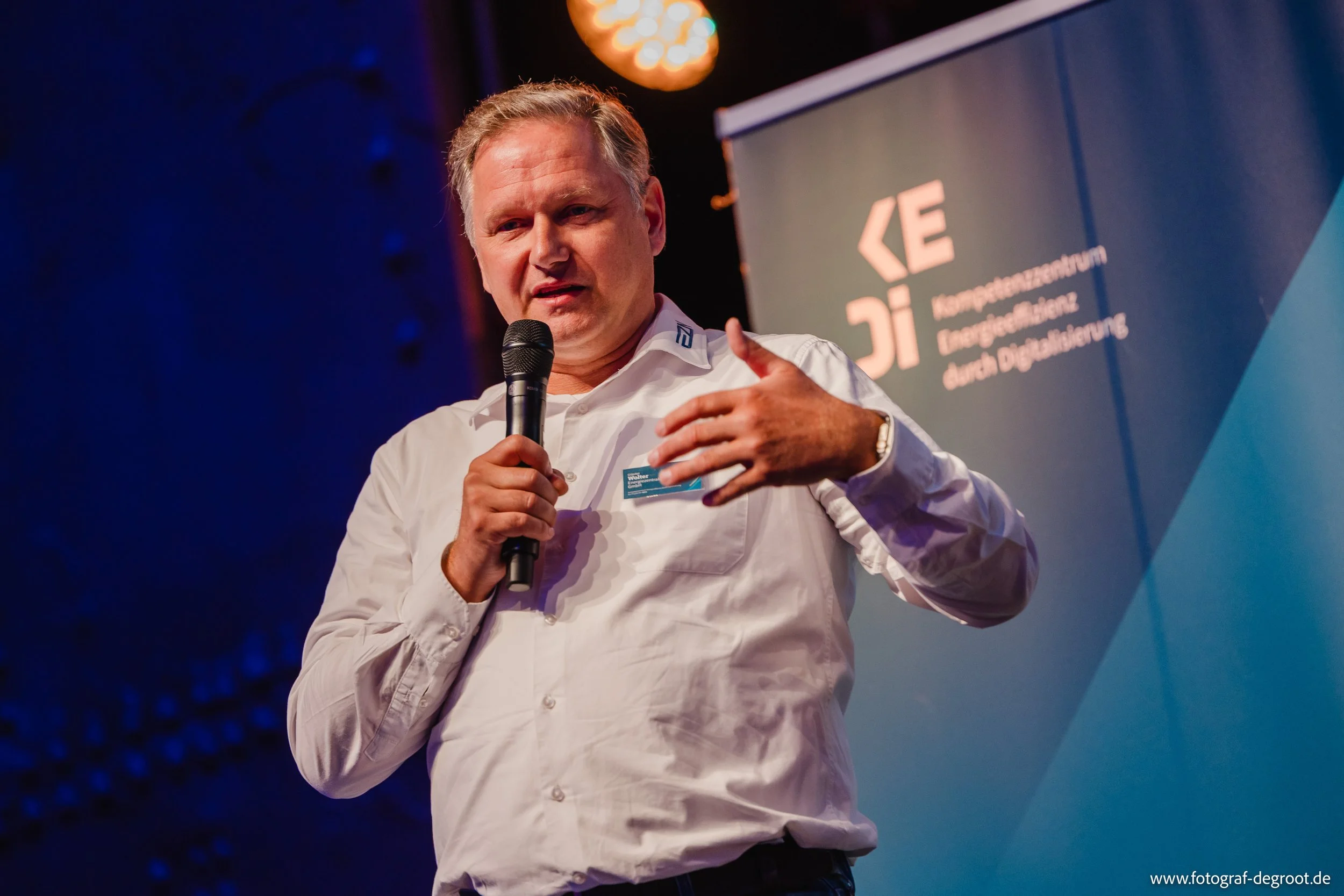 Ein Mann hält ein Mikrofon und spricht bei einer Veranstaltung. Im Hintergrund ist ein Banner mit dem Logo des K.E. Transformators zu sehen, welcher für Digitalisierung steht.