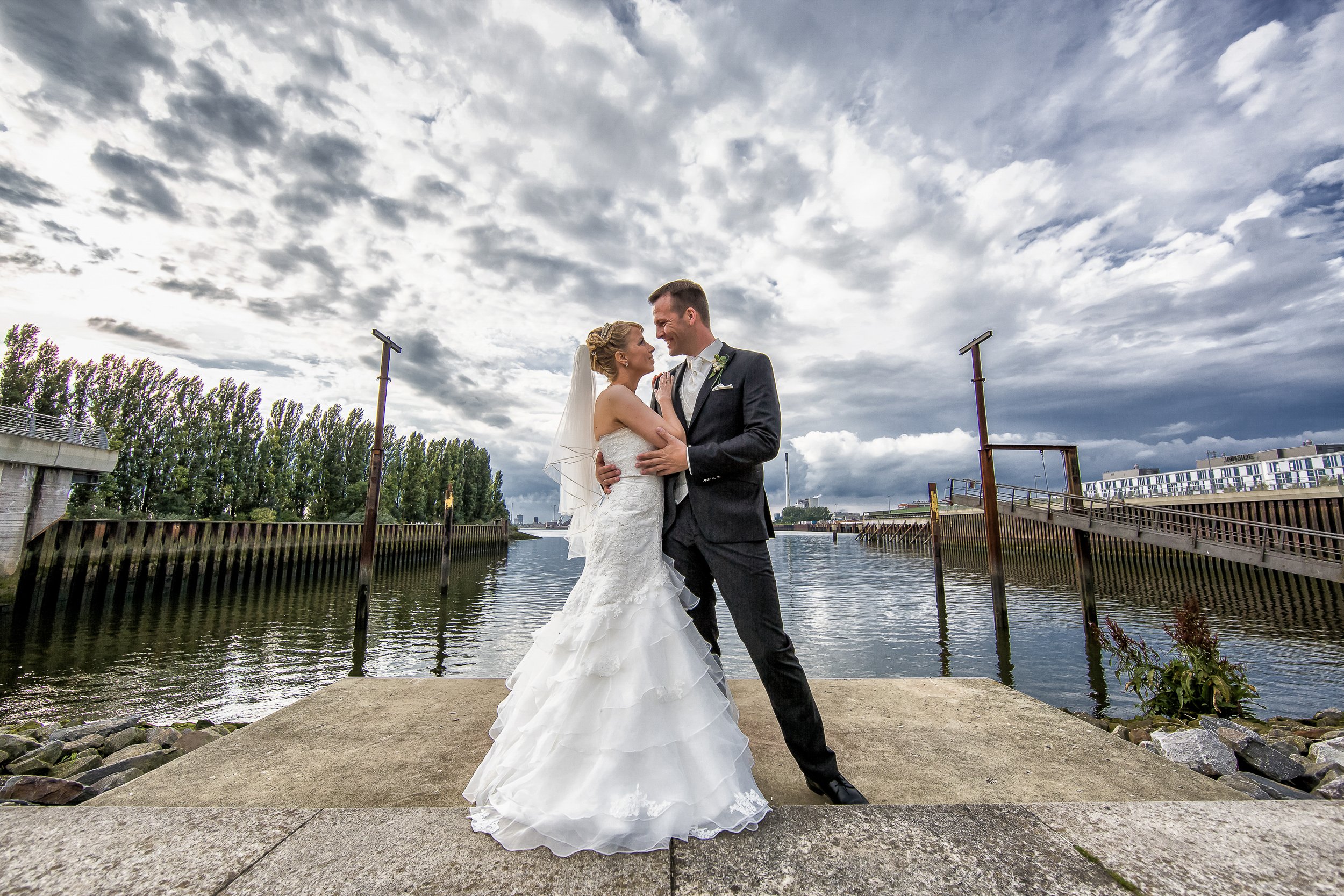 Ein Brautpaar bei ihrer Hochzeit steht am Wasser, umgeben von Wolken am Himmel, mit Bäumen im Hintergrund.