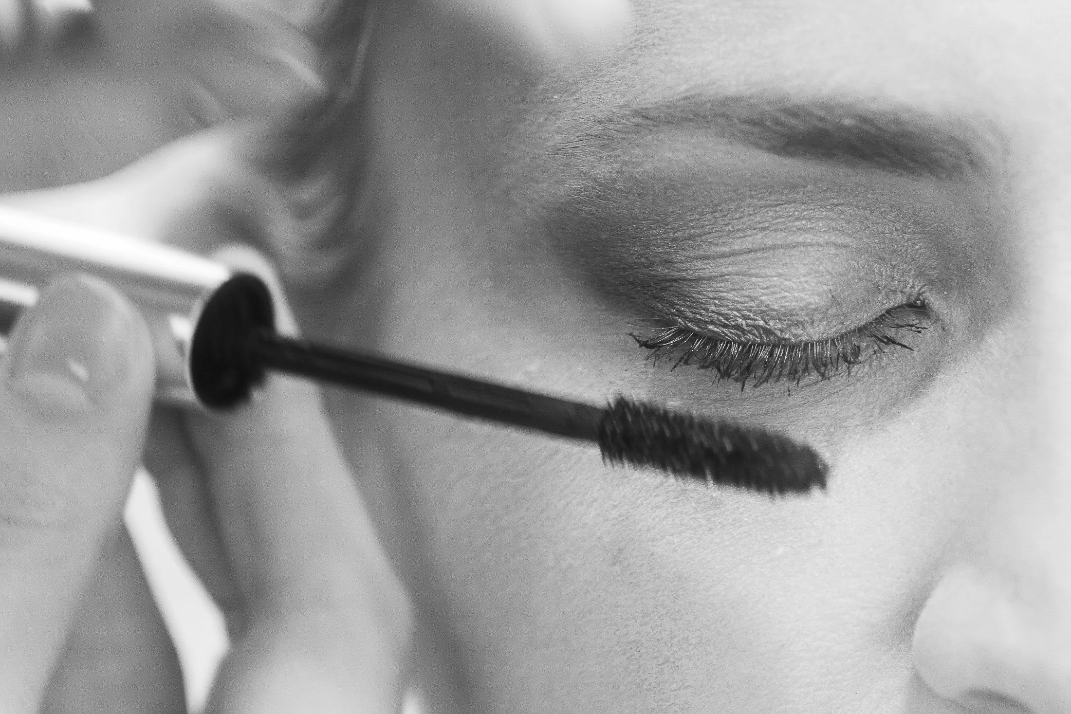Nahaufnahme eines geschlossenen Auges mit geschminkten Lashes, das auf einer glatten Haut liegt. Eine Person trägt eine Pipette, die vermutlich Produkt auf die Haut aufträgt.