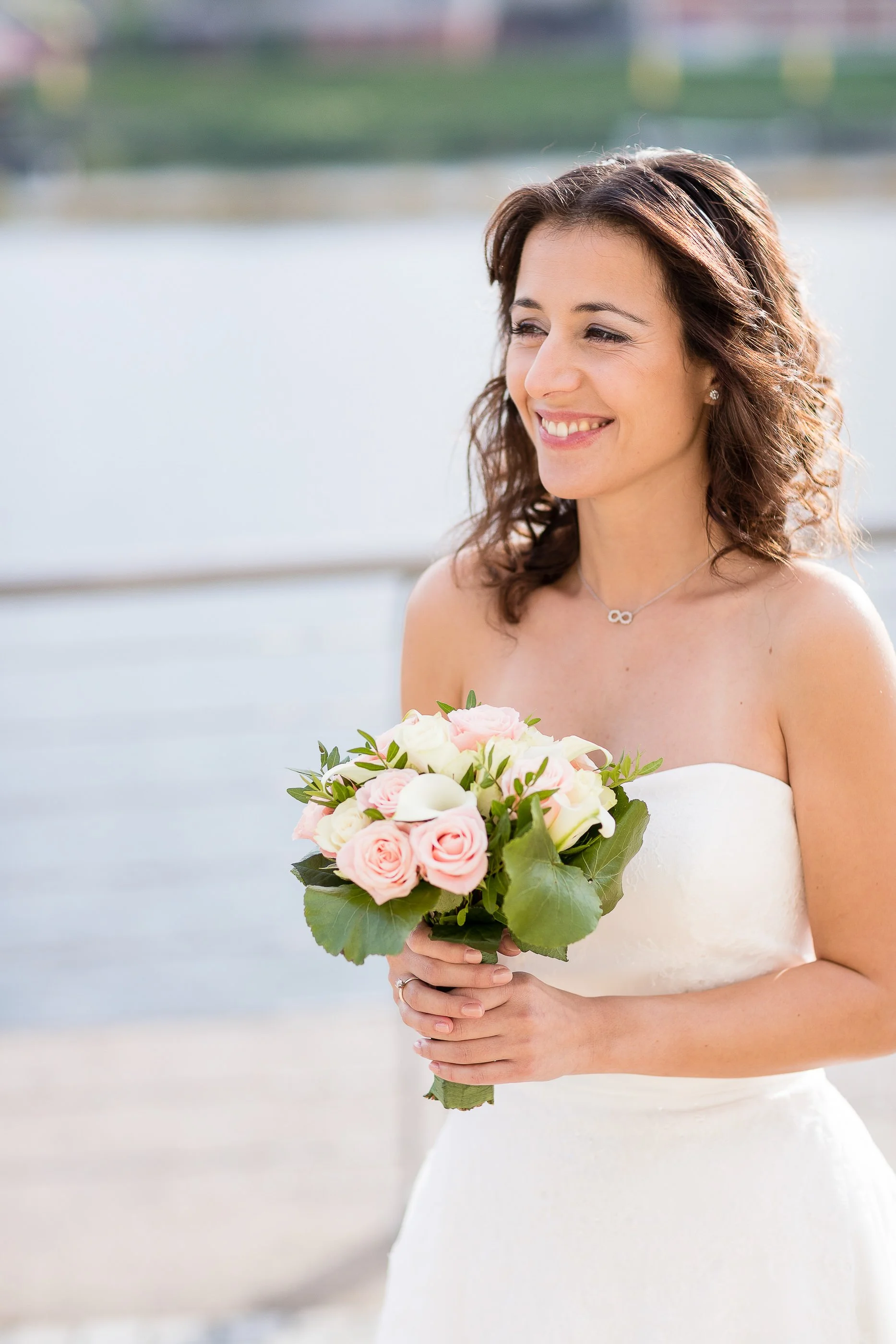 Eine lachende Frau in einem weißen Kleid hält einen Blumenstrauß mit rosa und weißen Blumen vor sich. Der Hintergrund zeigt Wasser und eine Stadtansicht bei Tageslicht.