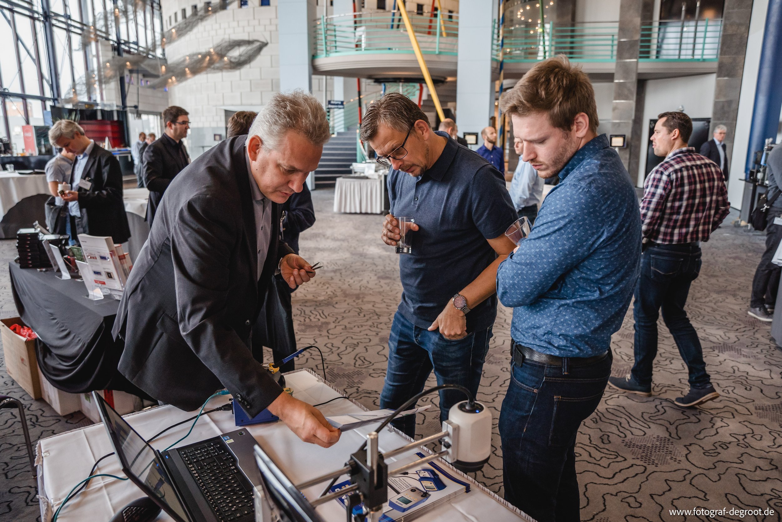 Menschen in einem modernen Konferenzraum, die sich um einen Tisch mit technischen Geräten und Computern versammeln. Eine Person zeigt auf etwas auf dem Tisch, während andere zuschauen.