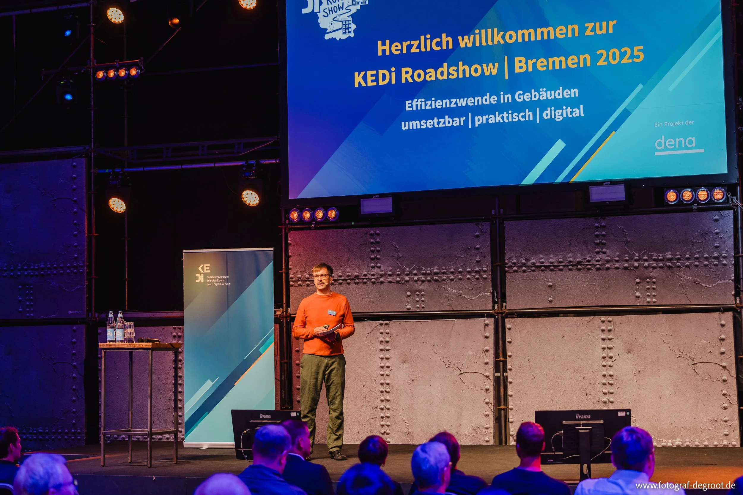 Ein Mann steht auf einer Bühne bei einer Präsentation, im Hintergrund ein großer Bildschirm mit dem Text 'Herzlich willkommen zur KEDI Roadshow Bremen 2025'. Im Publikum sitzen mehrere Personen und schauen zu. Es gibt auch einen Stehtisch mit Wasserf