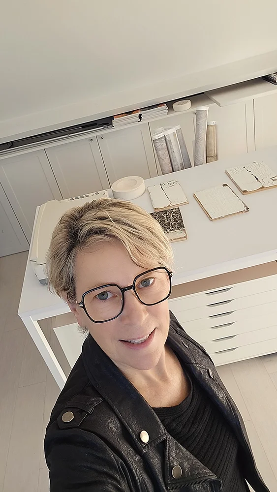 Selfie of a woman with short blonde hair and black round glasses, wearing a black leather jacket, standing in a room with a white table behind her on which there are several opened books or notebooks and a white device, with white cabinets and shelves in the background.