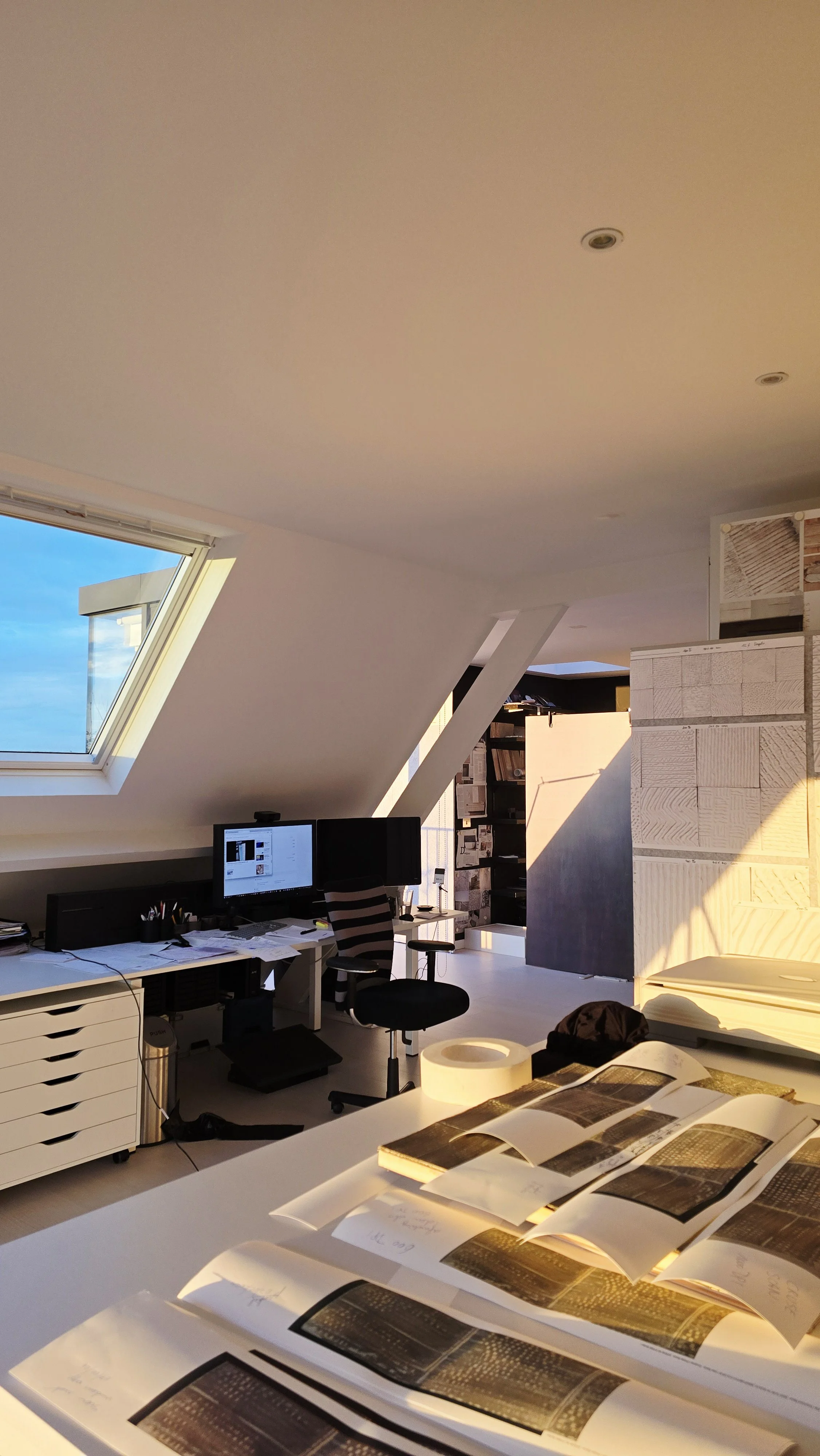 Photo of an office with a sloped ceiling, a window showing the sky, a desk with multiple computer monitors, a swivel chair, and various office supplies, with sunlight illuminating part of the space.