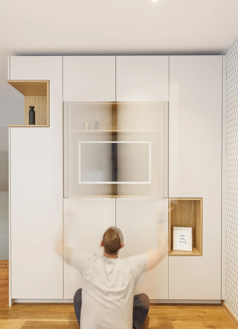 Ein Mann sitzt auf dem Boden und betrachtet eine weiße Wand mit eingebauten Schränken und offenen Regalen, in einem modernen, gut beleuchteten Raum.