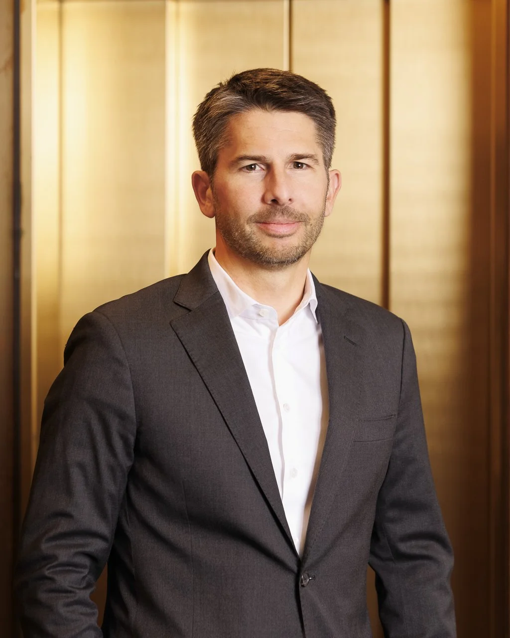 Ein Mann in einem schwarzen Anzug und weißem Hemd steht vor einer goldfarbenen Wand.