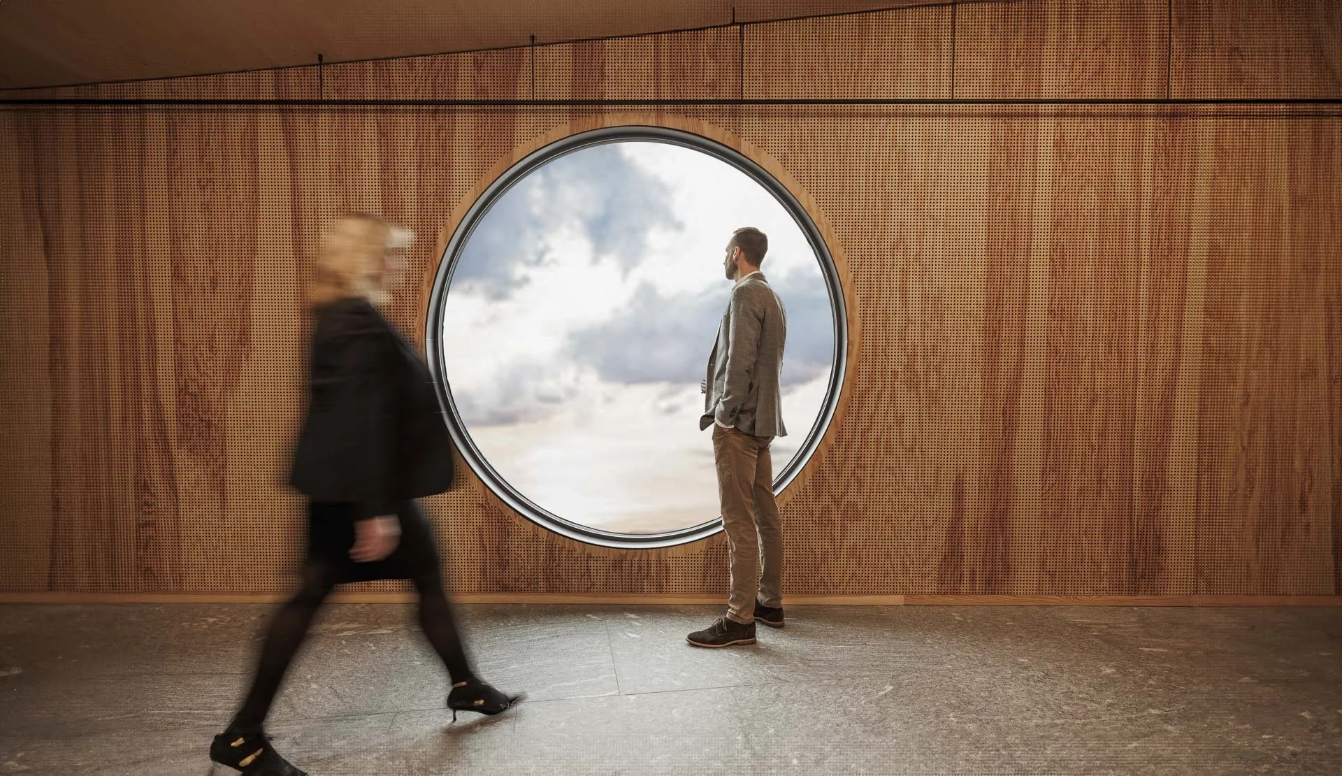 Ein Mann steht vor einem runden Fenster, durch das man den Himmel mit Wolken sieht. Ein blonder Frau ist im Vordergrund, die vorbei läuft.