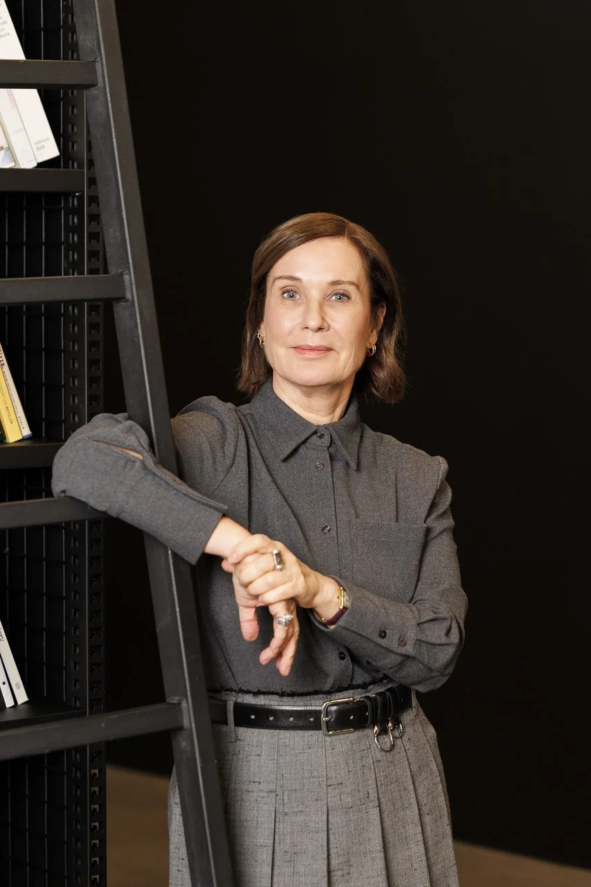 Eine Frau lehnt an einem schwarzen Bücherregal, trägt eine graue Bluse und einen schwarzen Gürtel, schaut zukunftsorientiert.