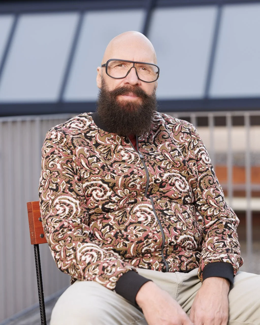 Ein Mann mit Glatze, Vollbart und Brille sitzt auf einem Stuhl auf einer Dachterrasse, trägt ein multicolores Muster-Sweatshirt und beige Hosen.