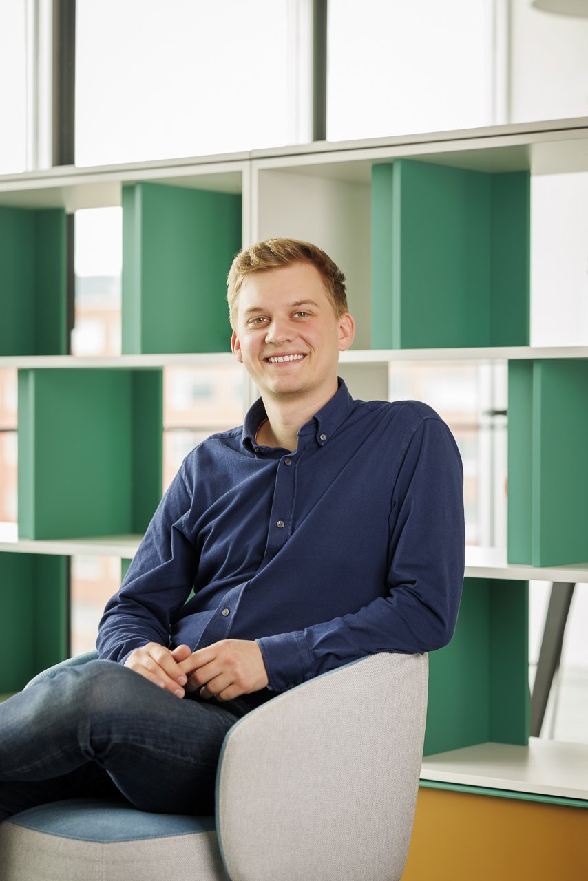 Ein junger Mann mit braunen Haaren sitzt in einem modernen Büro, trägt eine dunkelblaue Hemd und lächelt in die Kamera. Im Hintergrund grüne und weiße Regale.