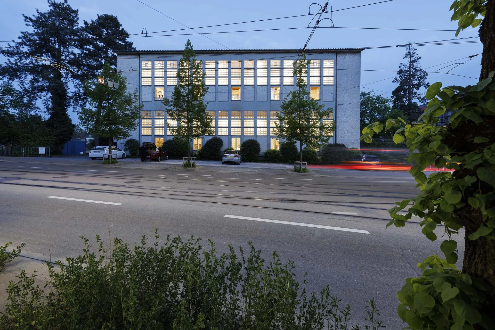 Aufnahme eines blauen mehrstöckigen Gebäudes bei Dämmerung mit erleuchteten Fenstern, umgeben von Bäumen. Vor dem Gebäude befindet sich eine Straße mit parkenden Autos.