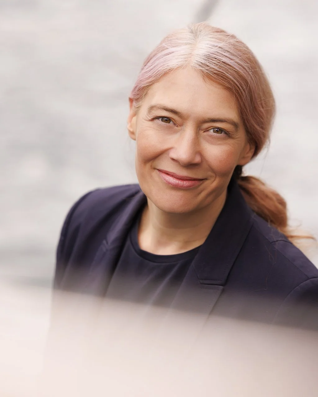 Porträt einer Frau mit hellbraunen, leicht rosa gefärbten Haaren, die über die Schulter fallen, lächelnd vor hellem Hintergrund.