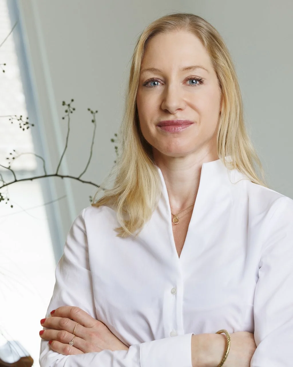 Eine Frau mit blonden, lockigen Haaren trägt ein weißes Hemd und blickt freundlich in die Kamera, während sie die Arme verschränkt.