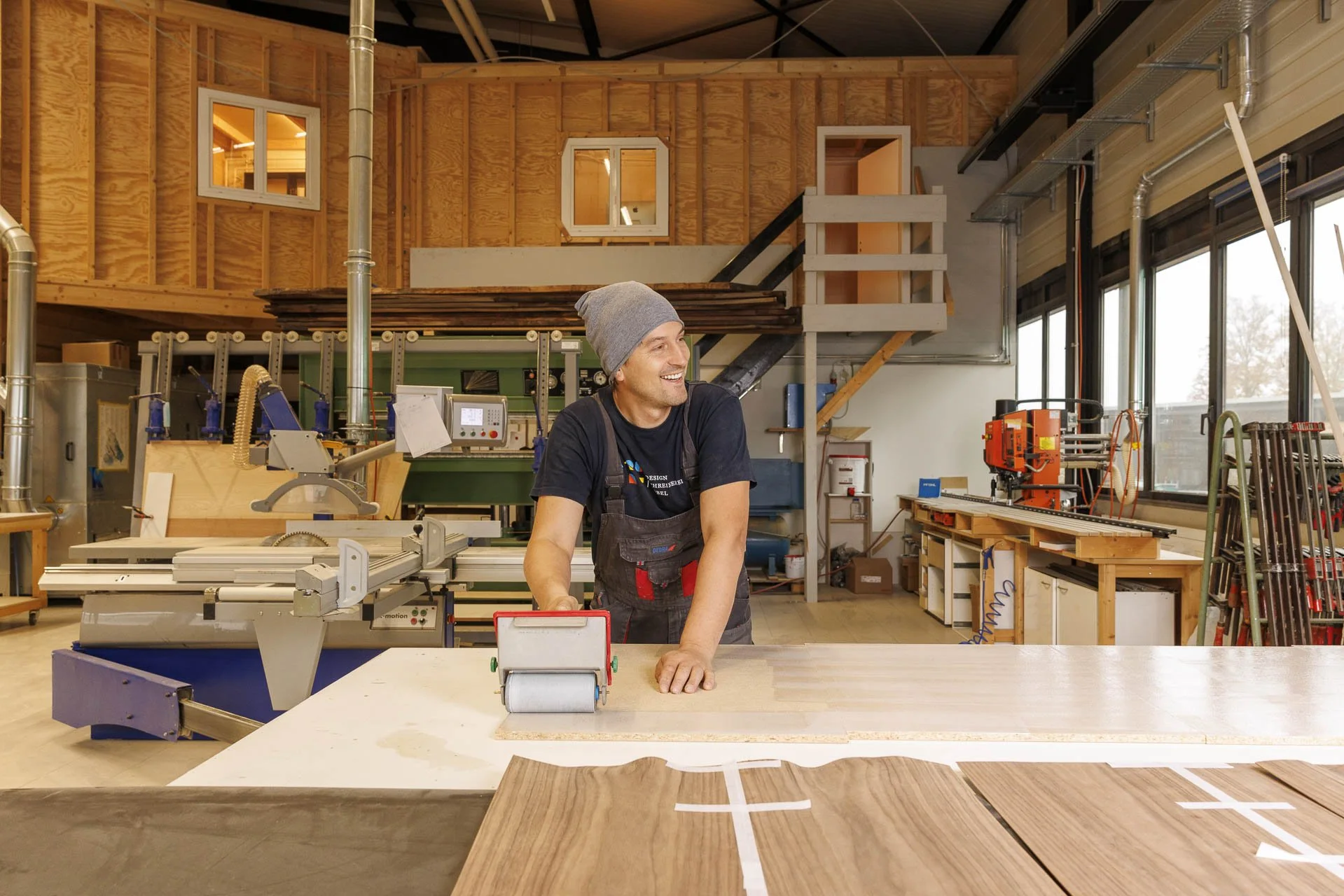 Schreiner arbeitet in einer Holzwerkstatt, trägt eine graue Mütze und eine schwarze Schürze, während er an einem großen Arbeitstisch steht.