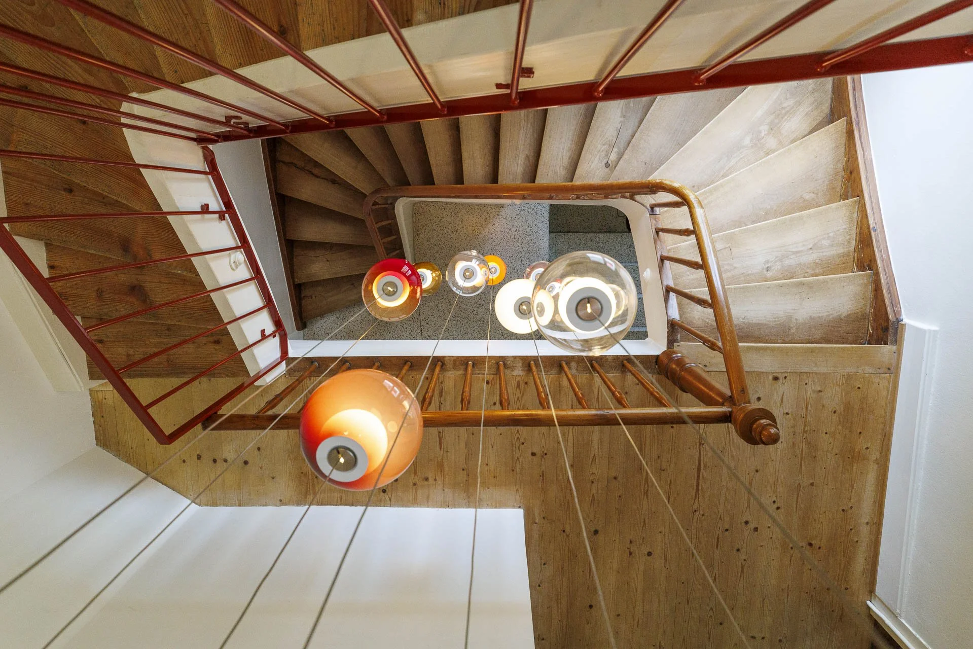 Blick nach oben auf eine mehrstufige Holztreppe mit roten Geländern, beleuchtet von mehreren dekorativen Hängeleuchten, die an Seilen hängen.