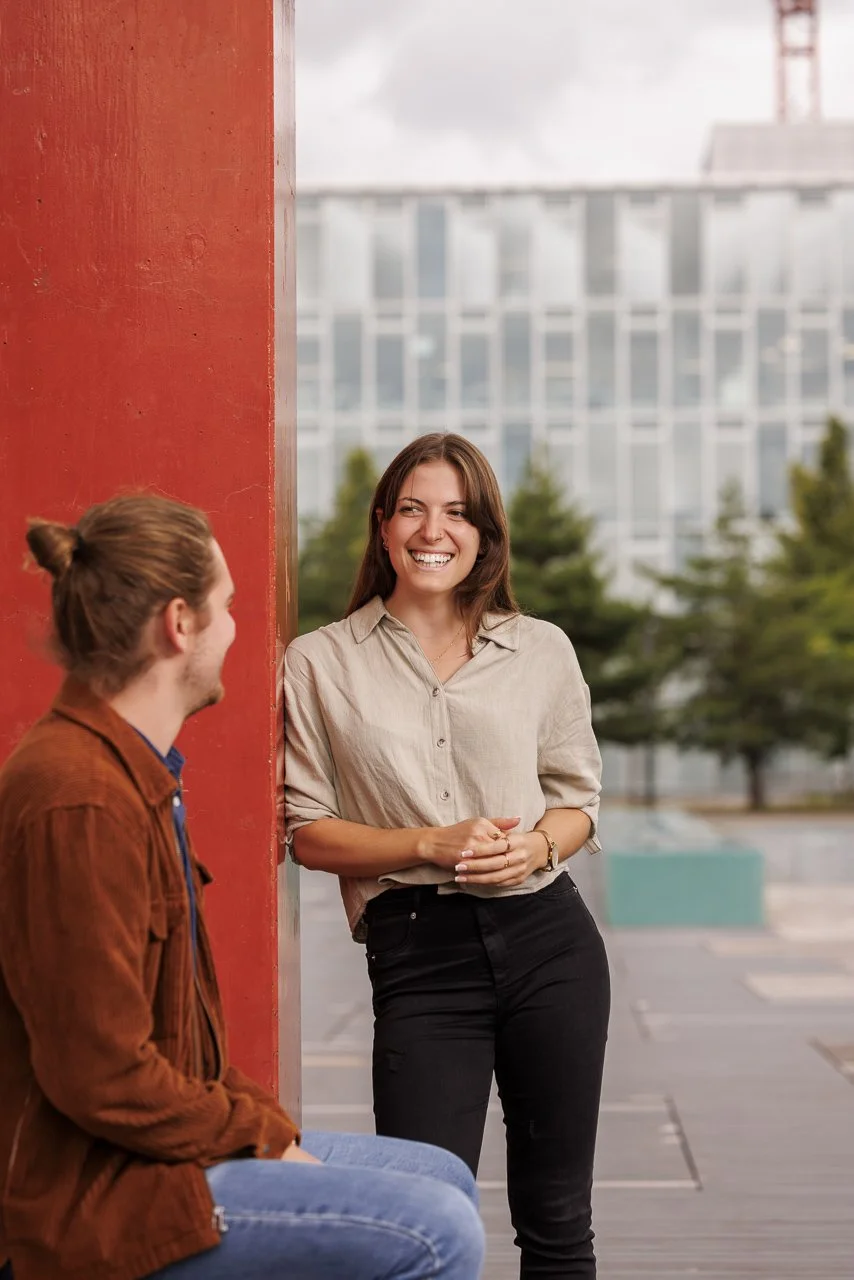 Zwei junge Menschen unterhalten sich im Freien, eine Frau steht lachend vor einem roten Wandstück, ein Mann sitzt mit seitlichem Blick zu ihr. Im Hintergrund sind moderne Gebäude und Bäume sichtbar.
