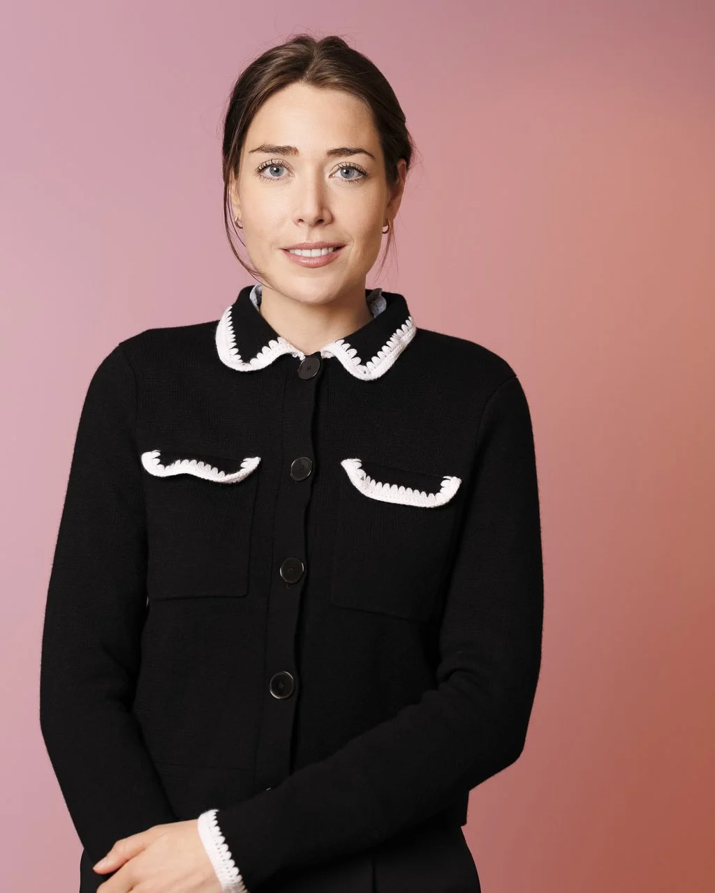 Ein Porträt einer Frau mit braunen Haaren, blauen Augen, trägt einen schwarzen Pullover mit weißen Ziernähten, vor einem pinken Hintergrund.