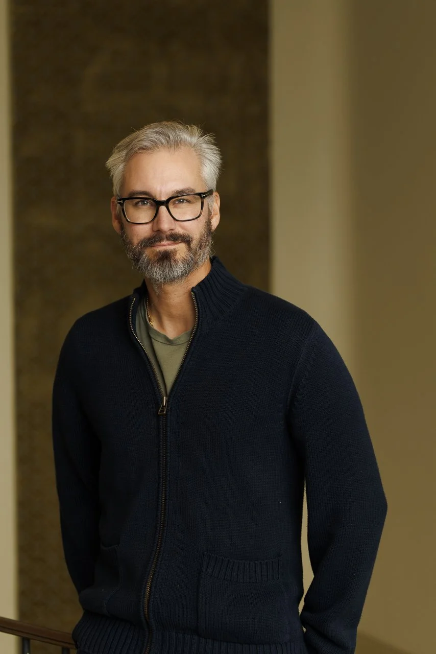 Ein Mann mit Brille, grau meliertem Haar und Bart trägt eine schwarze Jacke und steht in einem Innenraum.