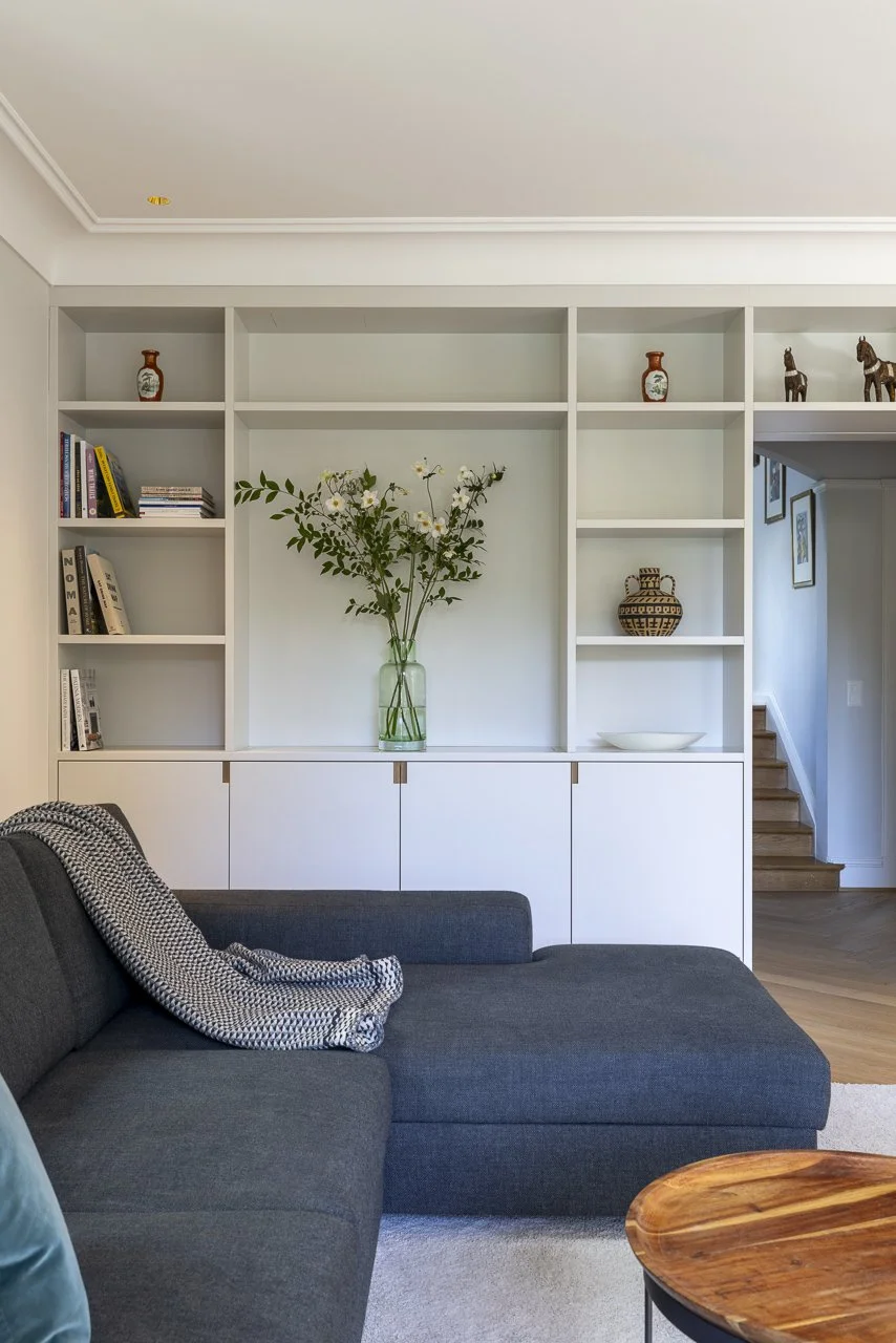 Wohnzimmer mit grauem Sofa, Holz-Couchtisch, weißem Bücherregal mit Vasen, Büchern und Dekorationsgegenständen, einer großen Vase mit Blumen und einer Treppe im Hintergrund.