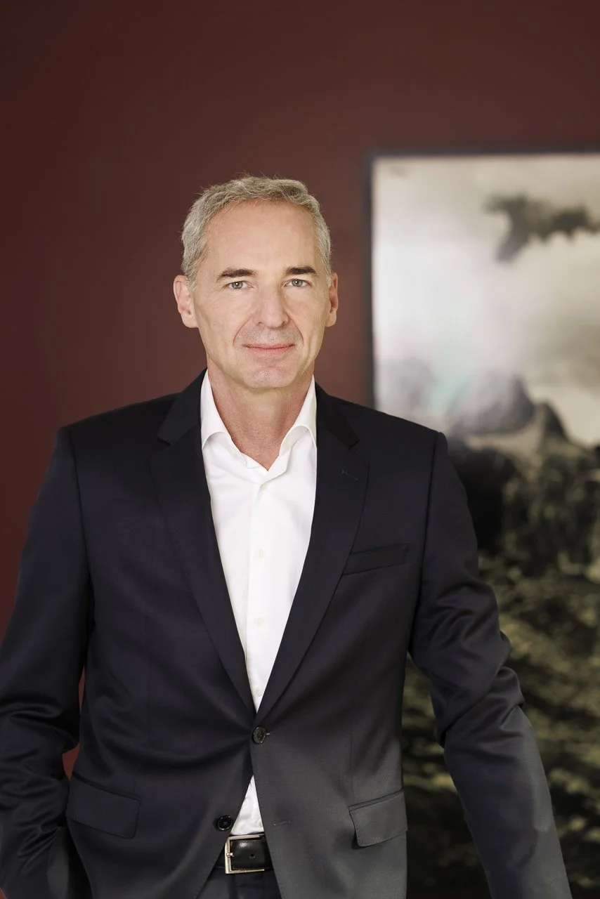 Ein Mann trägt einen schwarzen Anzug und ein weißes Hemd, steht vor einer Wand mit einem Kunstwerk im Hintergrund.