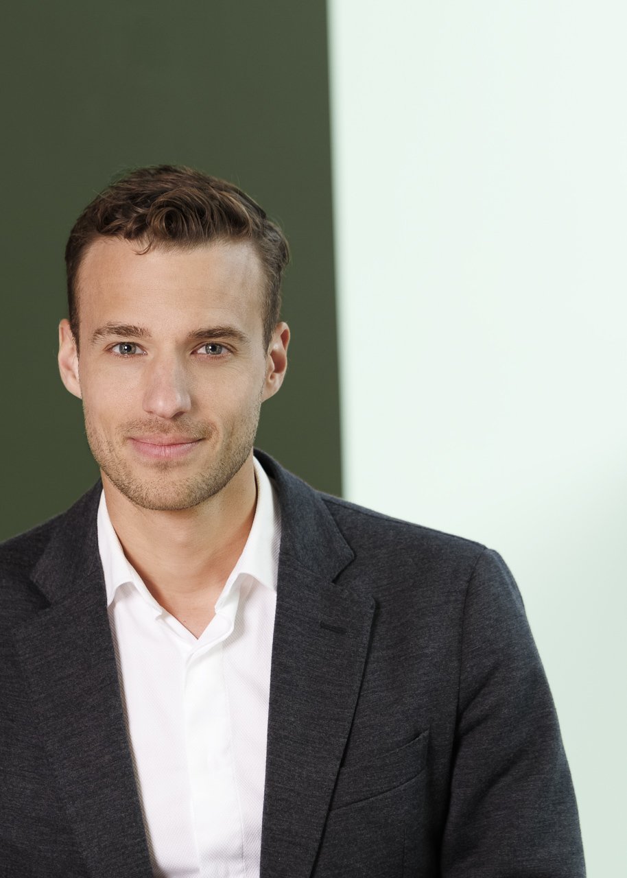 Ein junger Mann trägt einen dunklen Anzug und ein weißes Hemd, blickt freundlich in die Kamera, vor einem modernen, zweifarbigen Hintergrund in Grün- und Hellgrautönen.