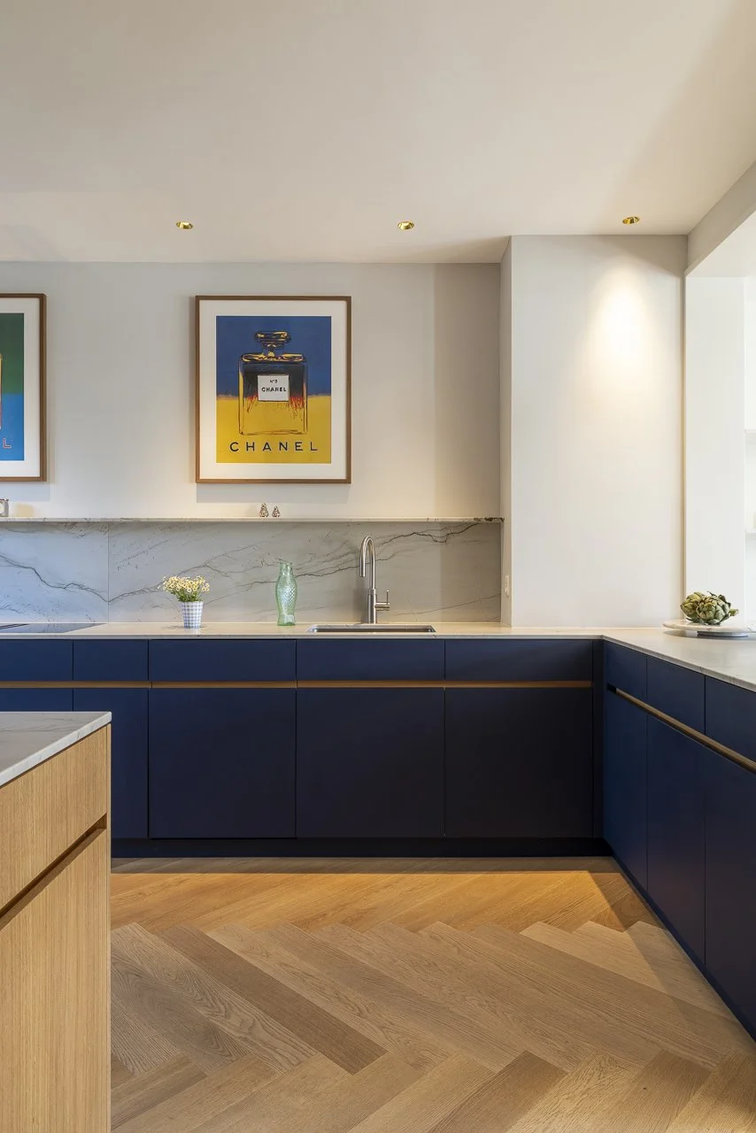 Moderne, minimalistische Küche mit blauen Schränken, weißer Marmor-Arbeitsplatte, Wandkunst von Chanel, kleiner Blumentopf, grünschimmernder Vasen und ein Fenster, das viel Tageslicht hereinlässt.