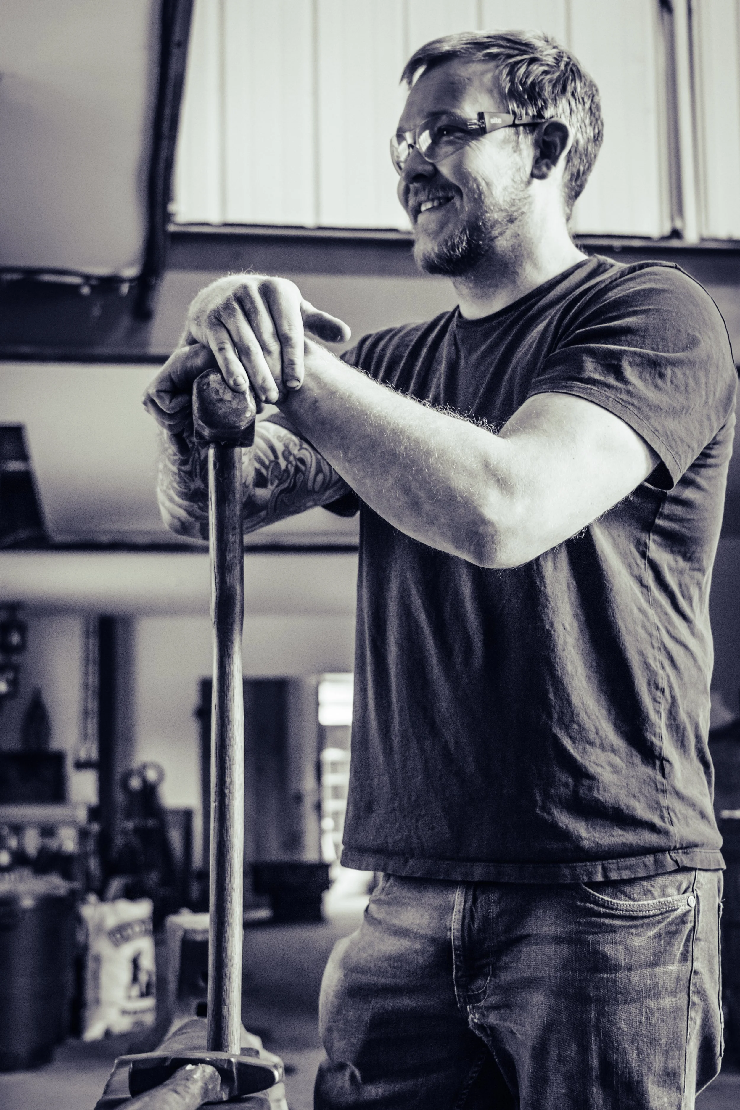 Dan, A blacksmith at IAStudios, in the forge holding a sledgehammer. He is smiling and looking to the side watching as his students work hard.