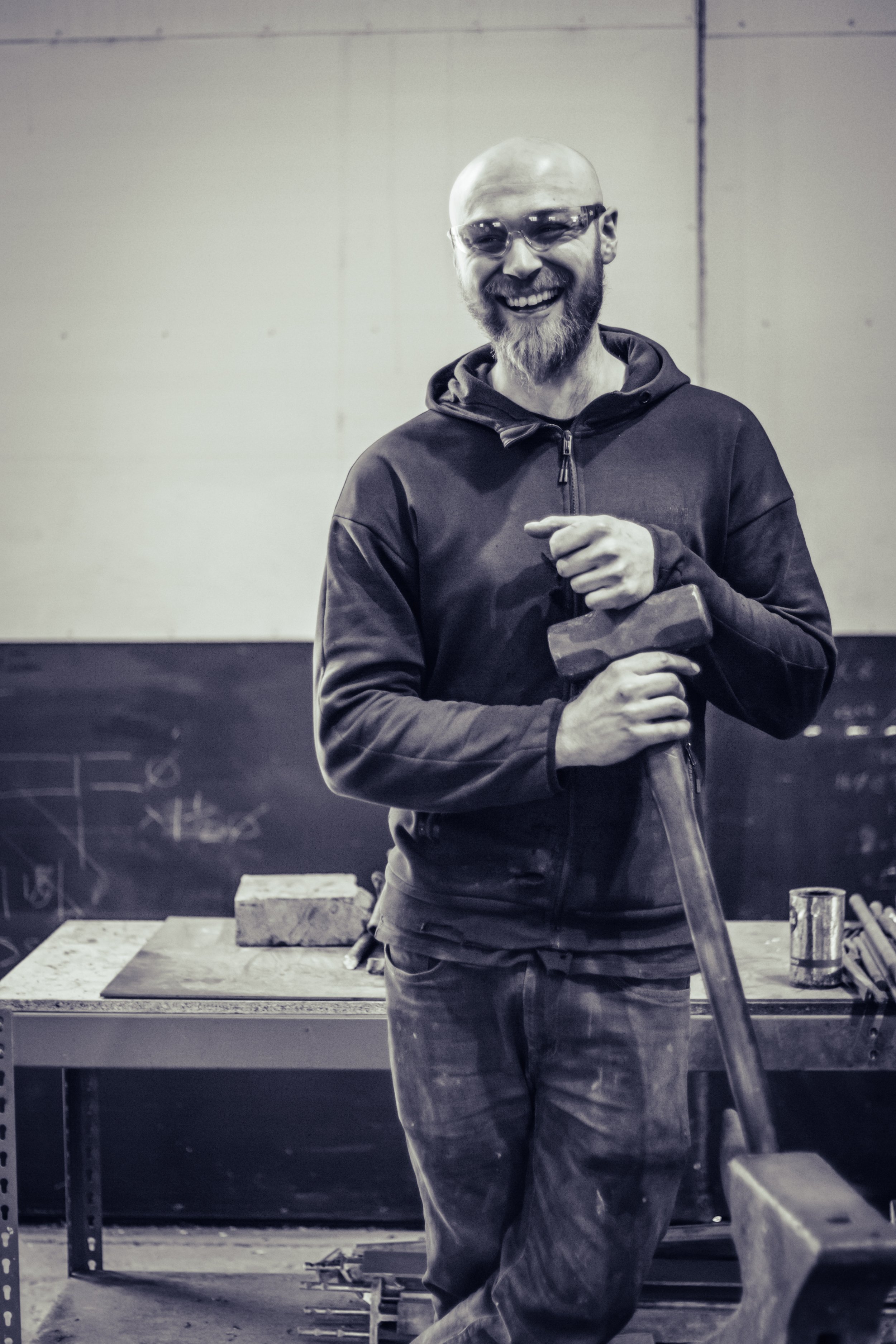 A bearded man with glasses smiling while holding a hammer in a workshop with a blackboard and tools in the background.