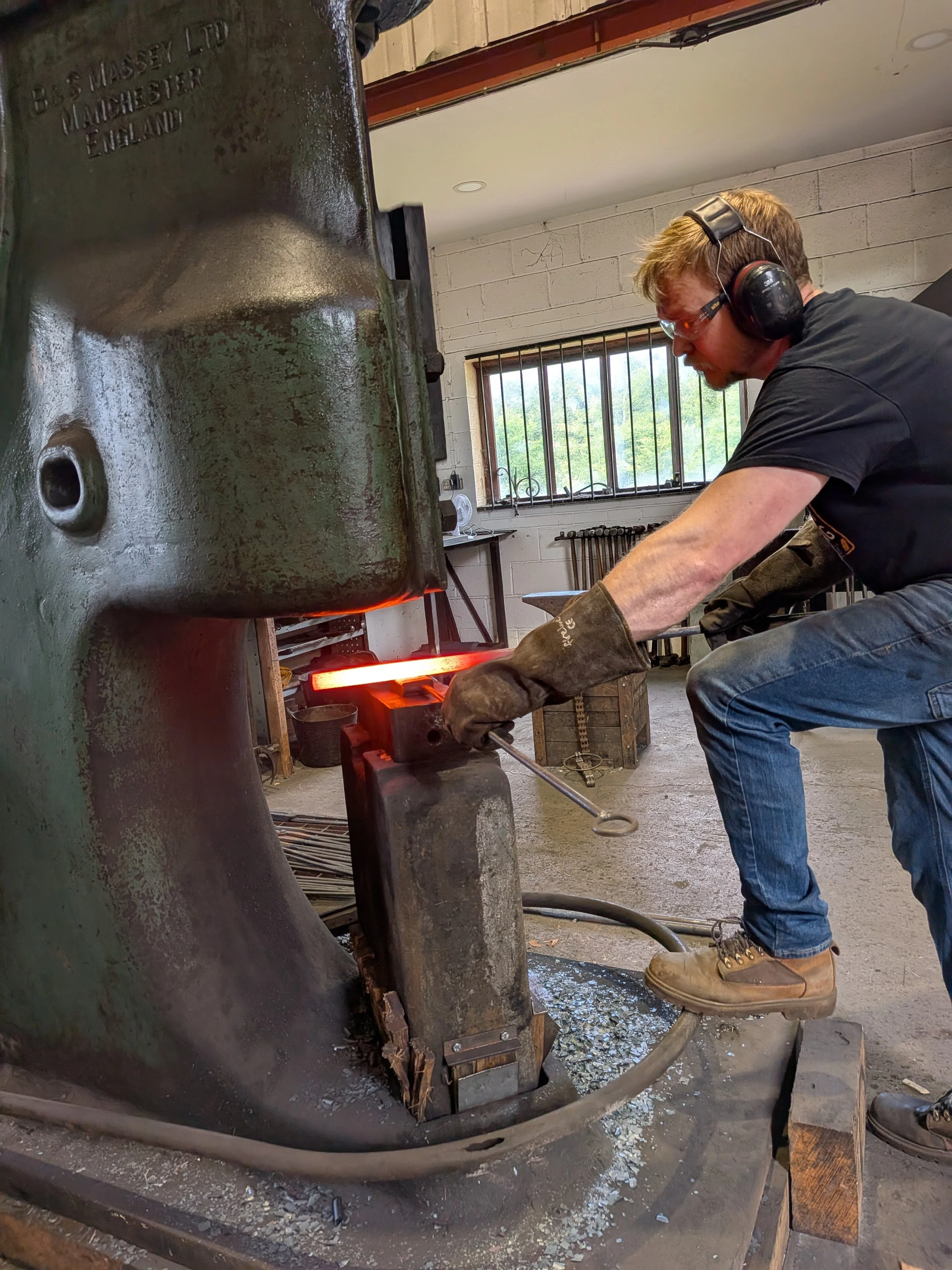 A blacksmith forging a bar of steel into a pair of tongs using a power hammer