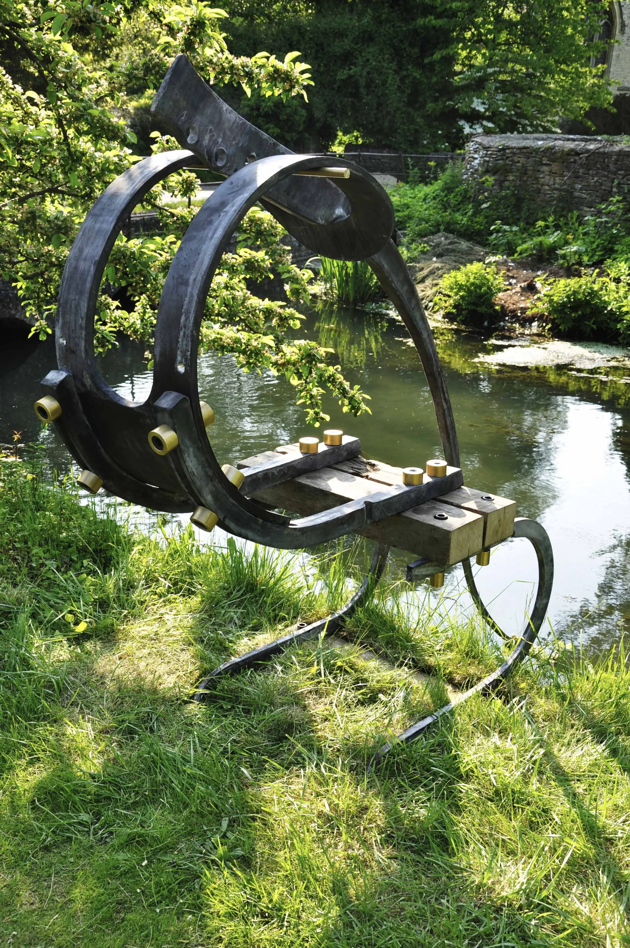 Sculpture of a wheelbarrow made from metal and wood by a pond surrounded by greenery.