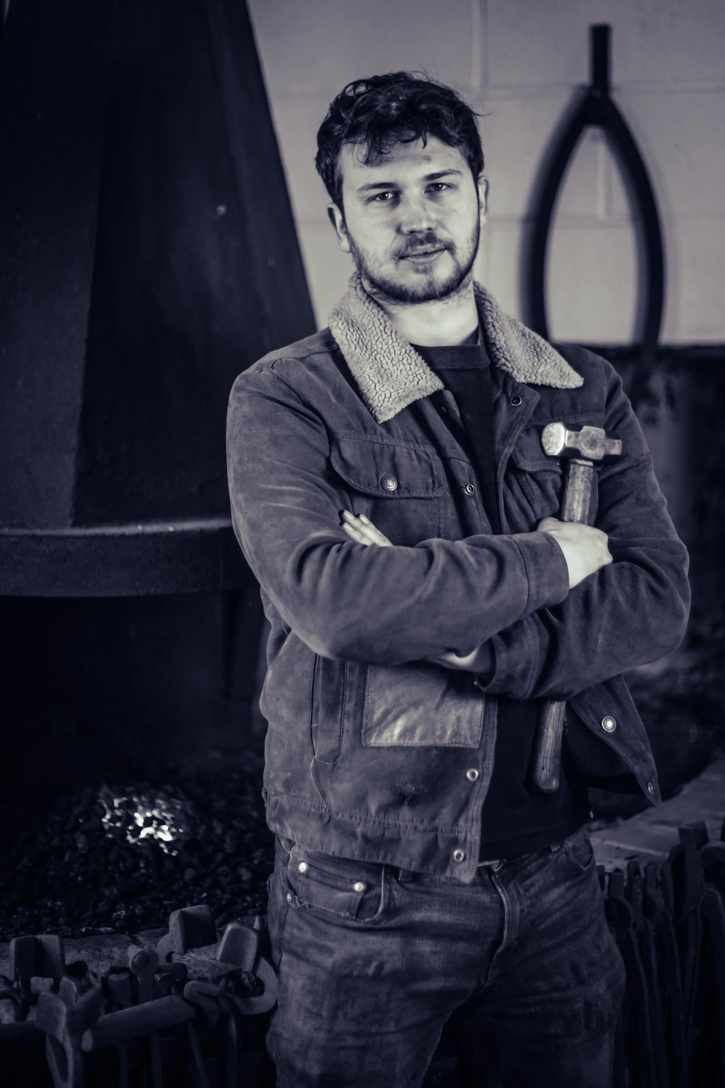 A young man with dark curly hair and a beard, wearing a jacket with a shearling collar, standing indoors with his arms crossed, holding a hammer, in front of a wall with a large black object in the background.
