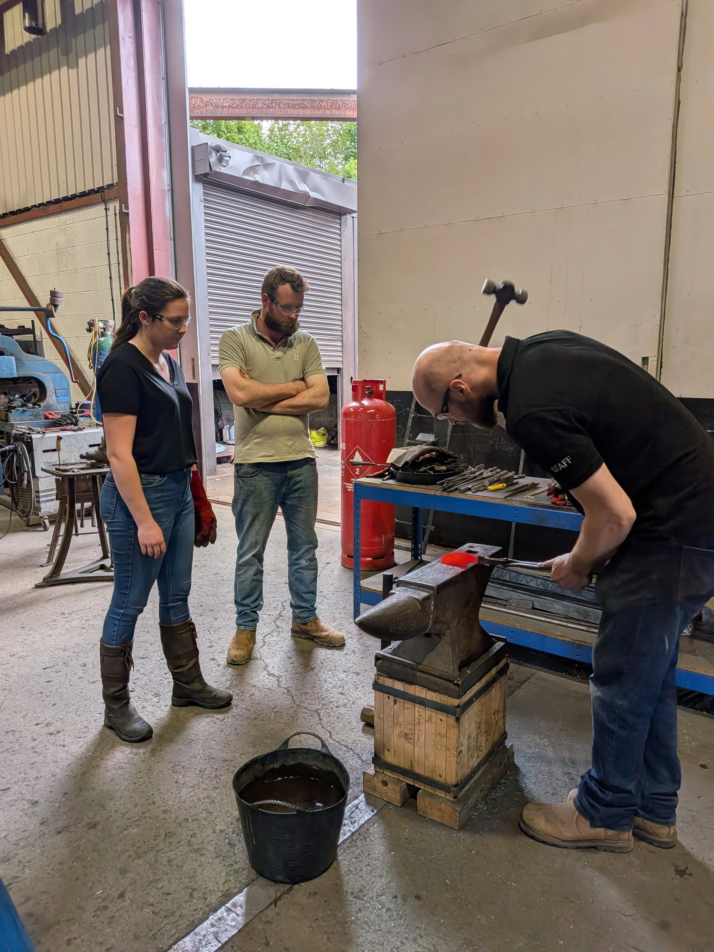 A blacksmith teaching students how to forge a knife. The blacksmith is swinging his hammer at a hot piece of steel as the students watch.