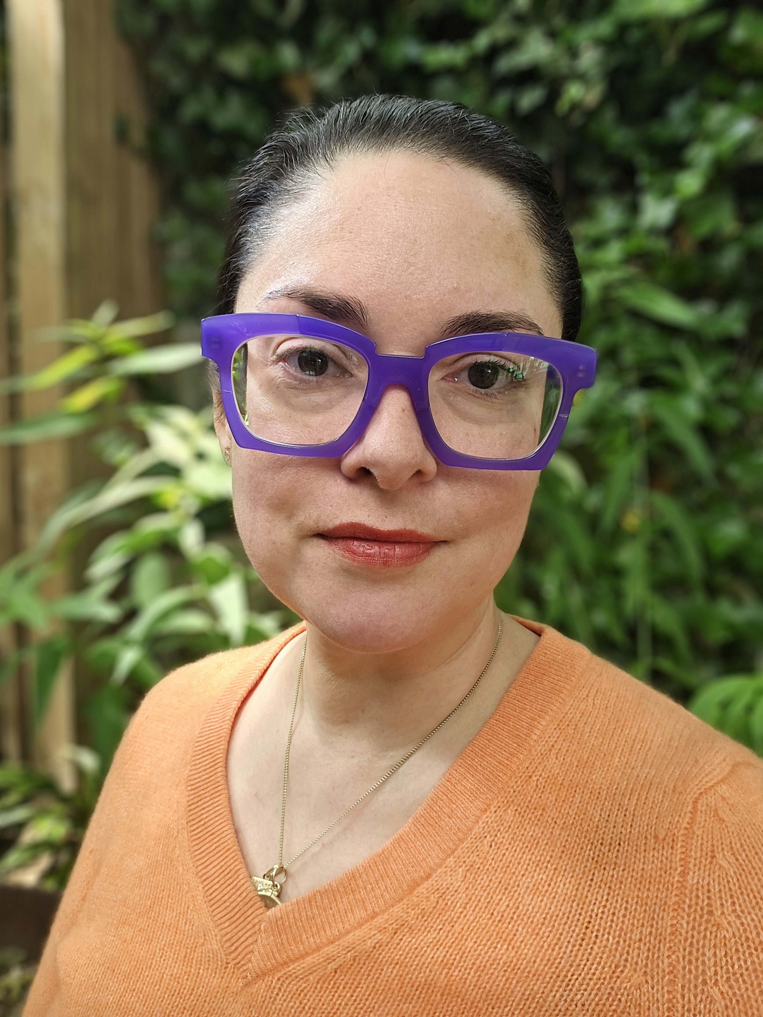 Close-up of a woman with dark hair, wearing purple glasses and an orange sweater, standing outdoors with green foliage in the background.
