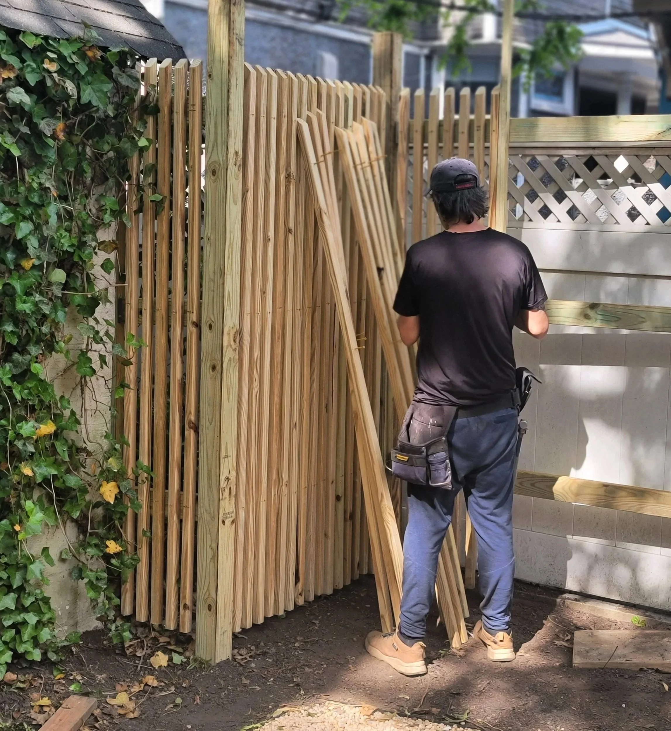 A person in a black t-shirt, gray pants, beige work boots, and a black cap is installing a wooden fence in a backyard. The fence panels are leaning against the posts they are securing.
