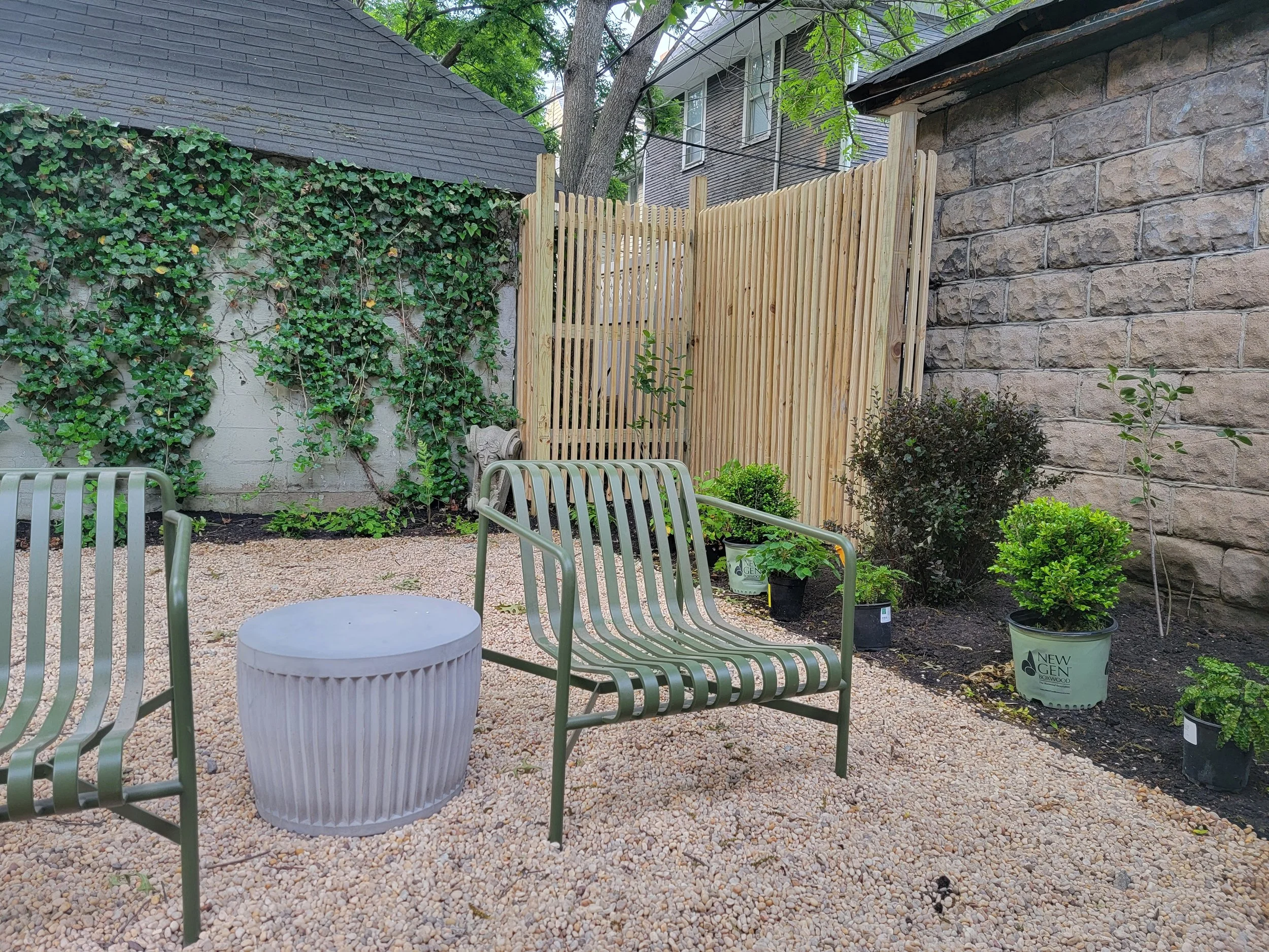A backyard patio with chairs, potted plants, a wooden fence, a stone wall, and a tree, with neighboring houses in the background.