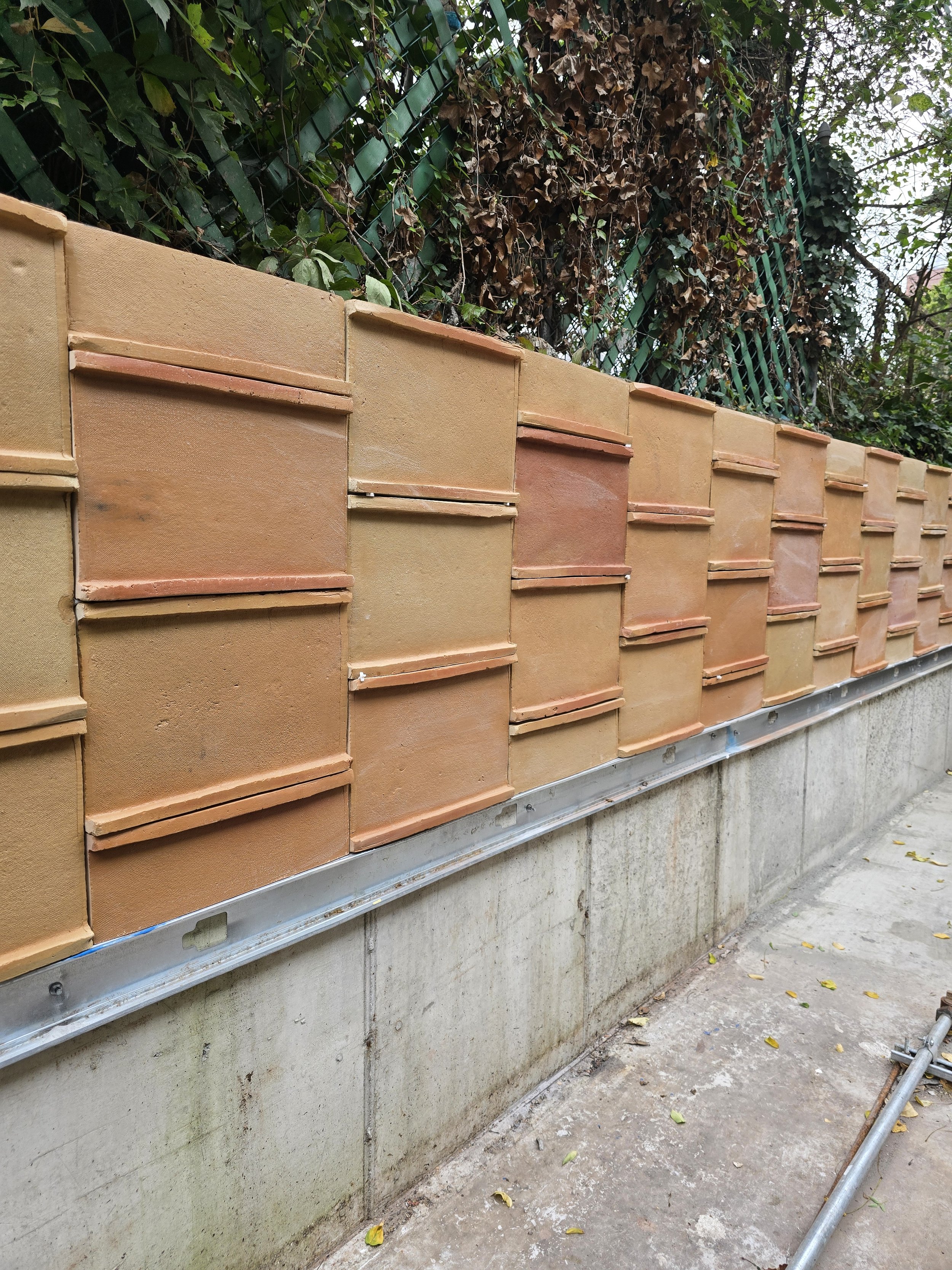 A retaining wall with clad with terracotta tiles. The wall is supported by a concrete plinth covered with fallen leaves, and there is a green fence with overgrown vines and foliage behind the wall.