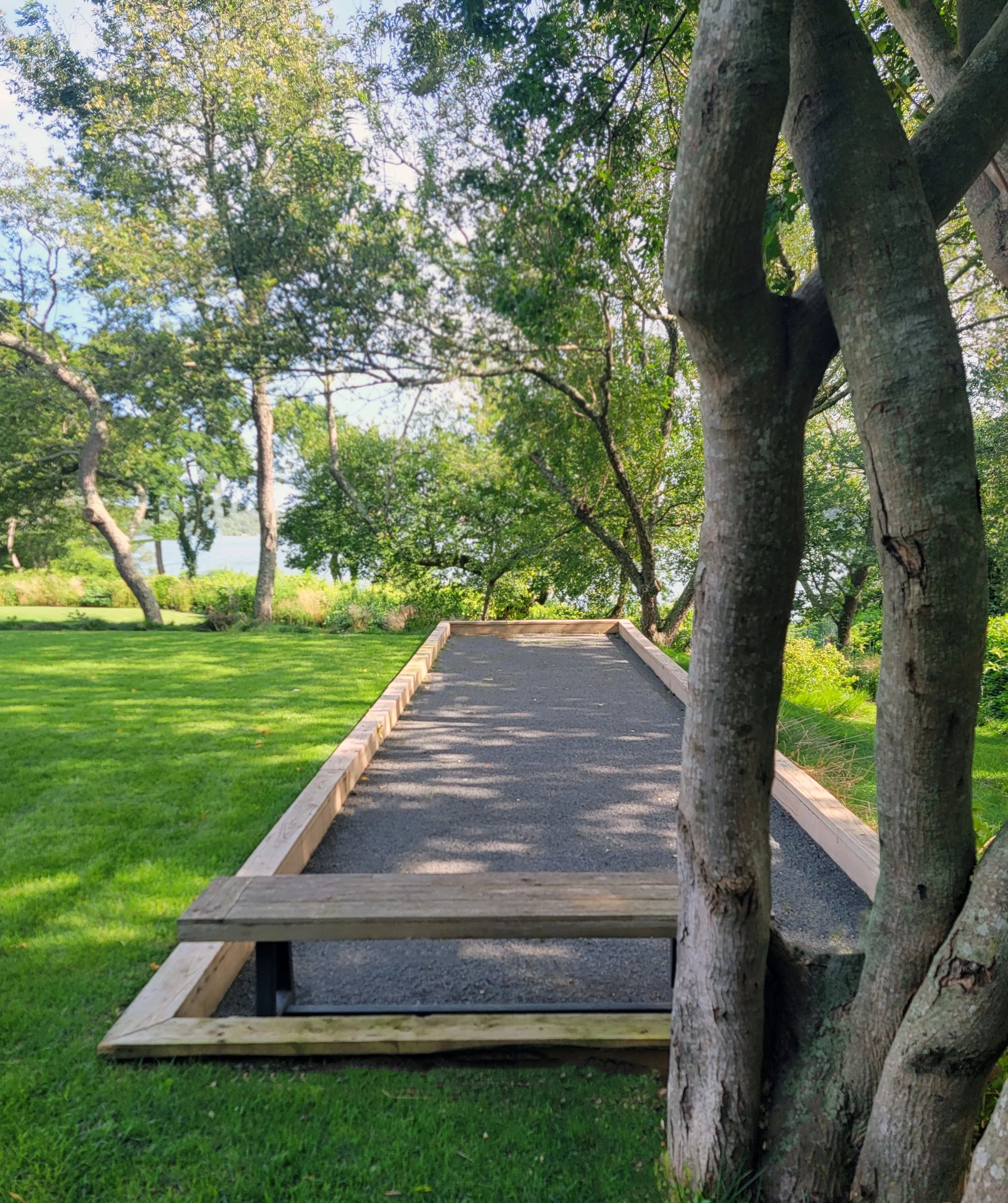 A gravel bocce court with wood edging and a bench, surrounded by grass and trees with green leaves, in a large residential backyard.