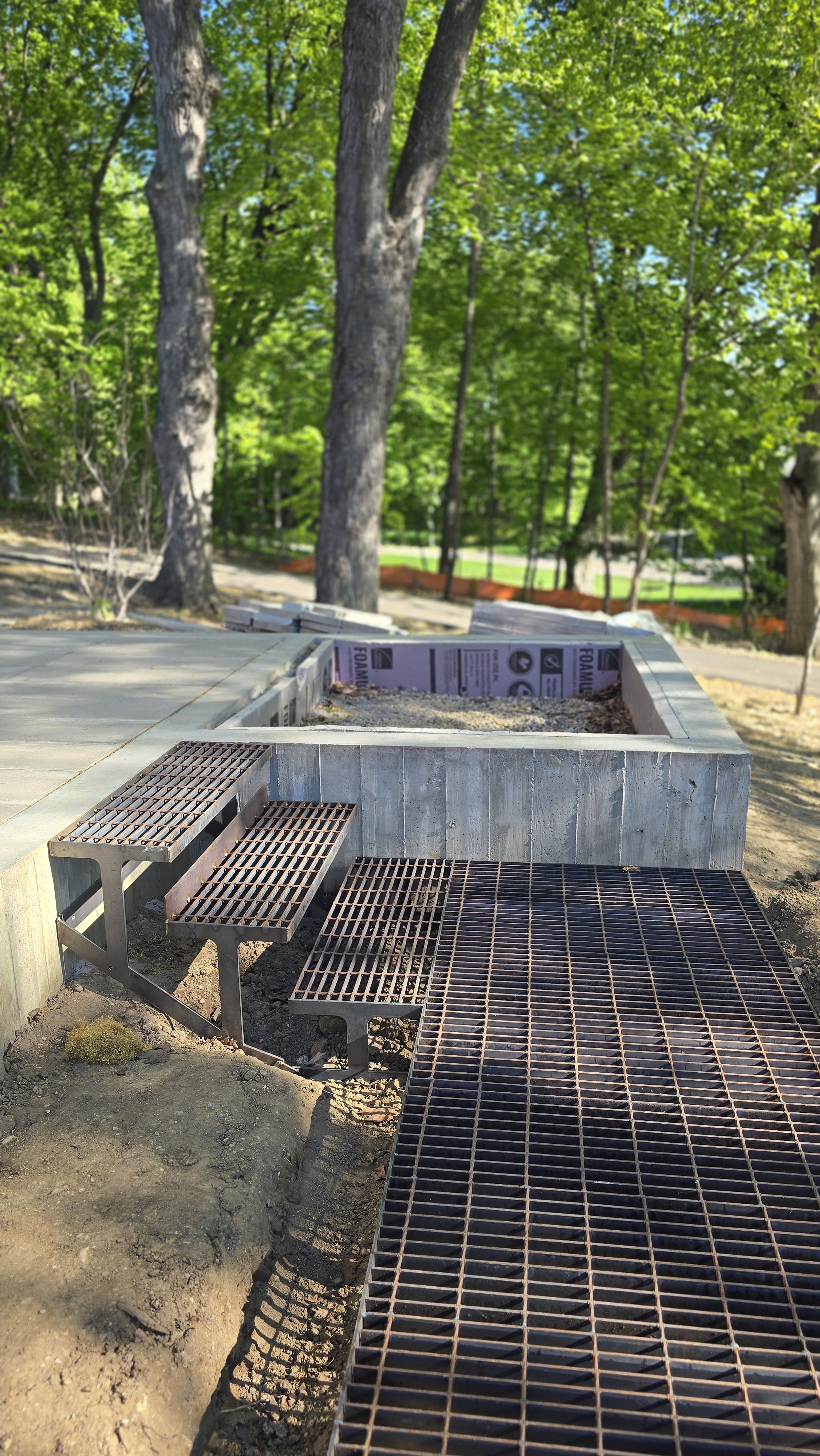 Construction site with metal grate walkway and steps, foundation bed with drainage boards in a green, wooded area.