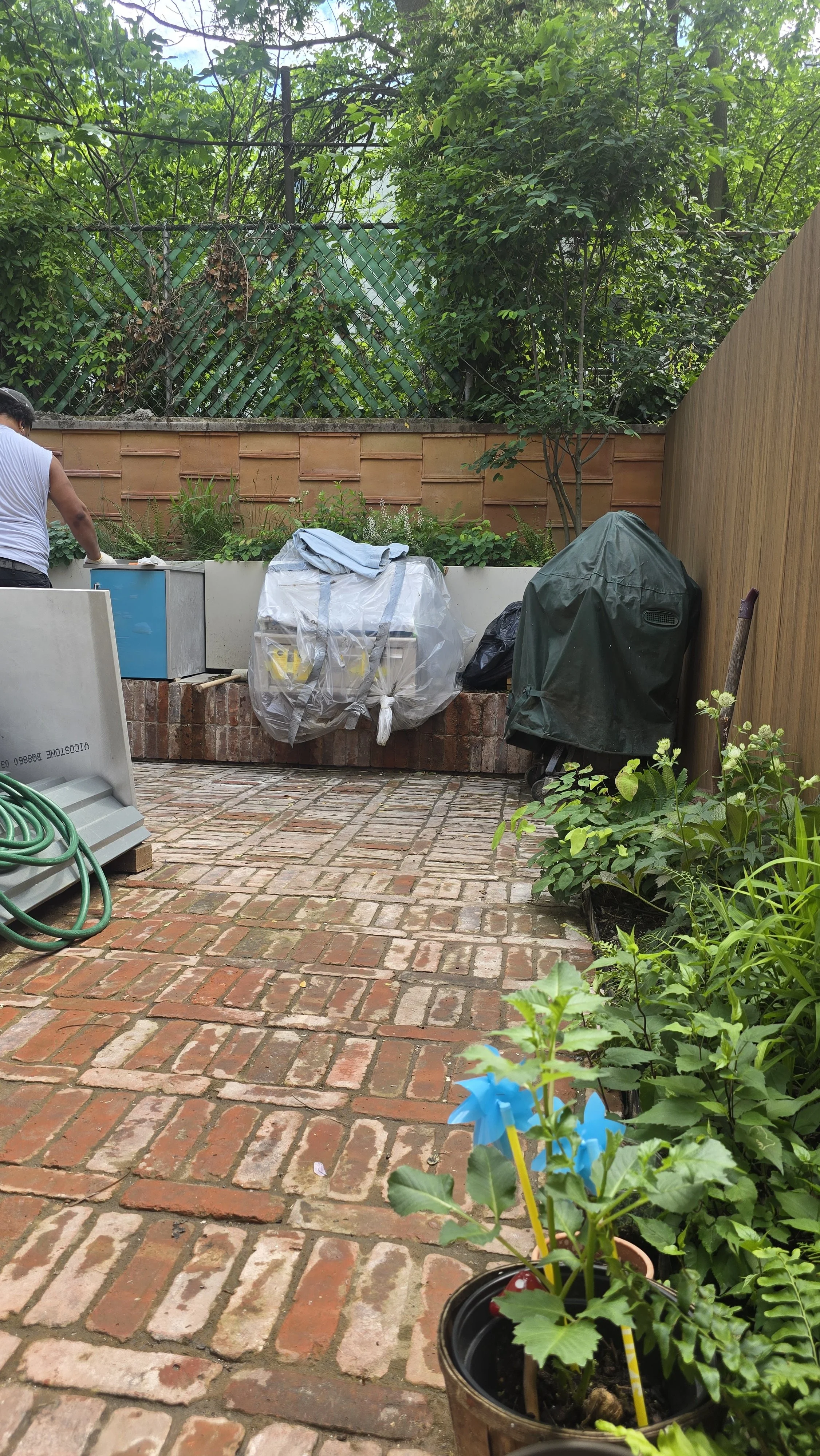 A backyard with a brick patio and garden beds, some covered or with tools, green plants, trees, and a wooden fence. A person is working on the left side, and there are gardening supplies, including a pot with a young plant in the foreground with blue