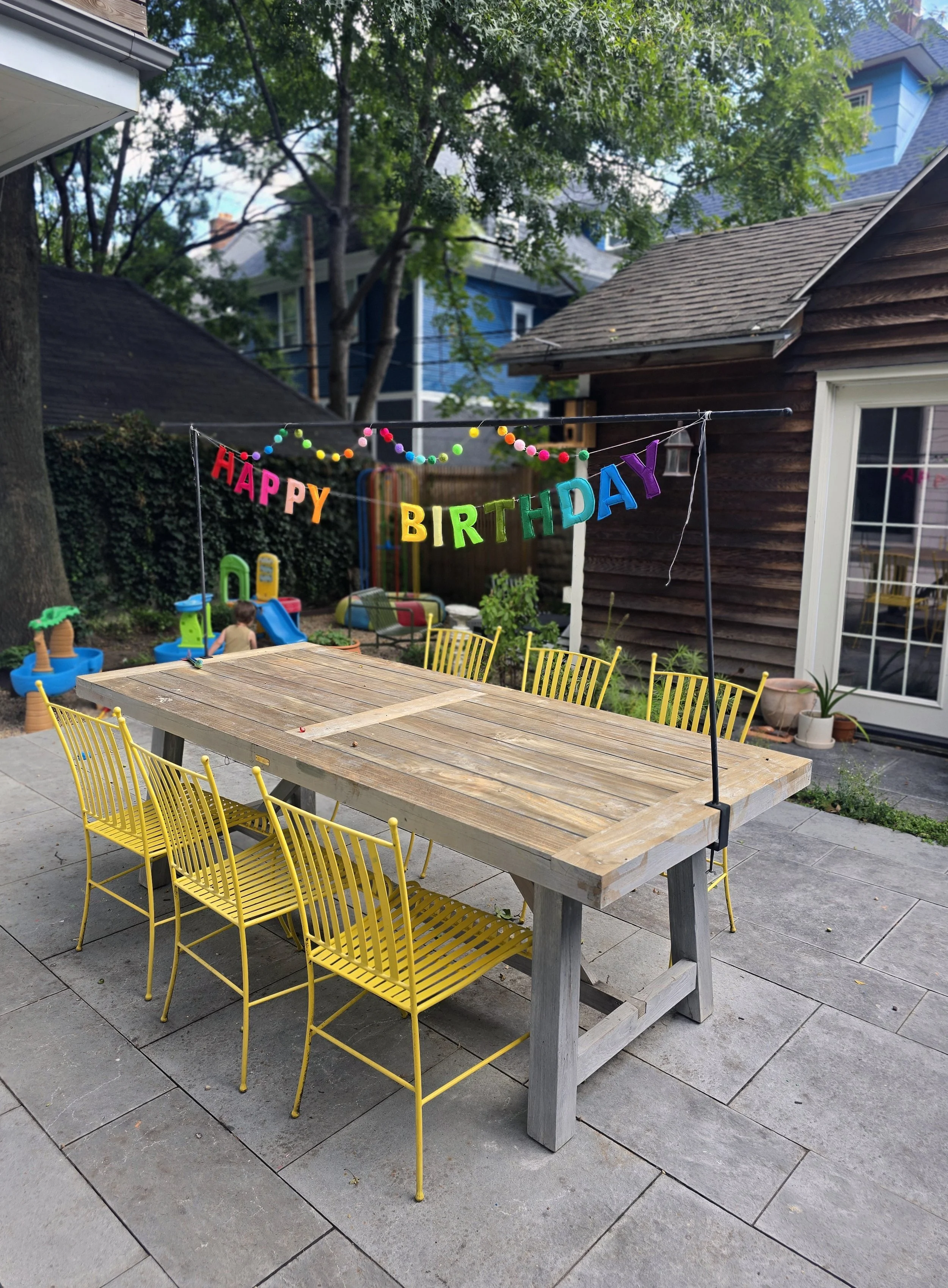 A backyard patio with a long wooden table surrounded by yellow chairs, decorated with a colorful 'Happy Birthday' banner and string lights, in front of a house with a wooden exterior and French doors.