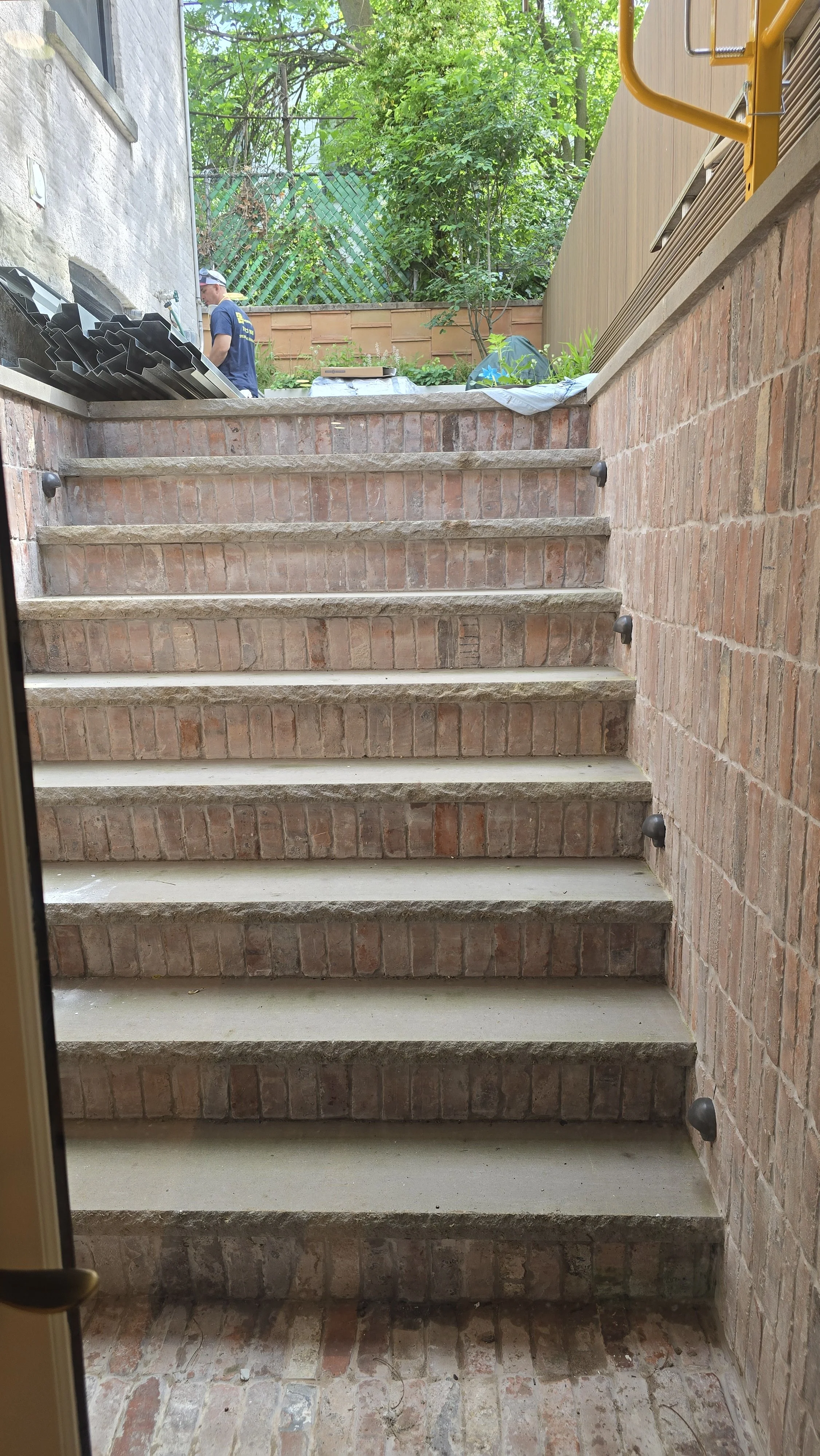 View of brick stairs leading outdoors, with a brick and cement floor at the bottom, surrounded by brick and wood-sided walls, and a small tree or shrub in the background. There is a person in a blue shirt and white cap standing near a pile of black m