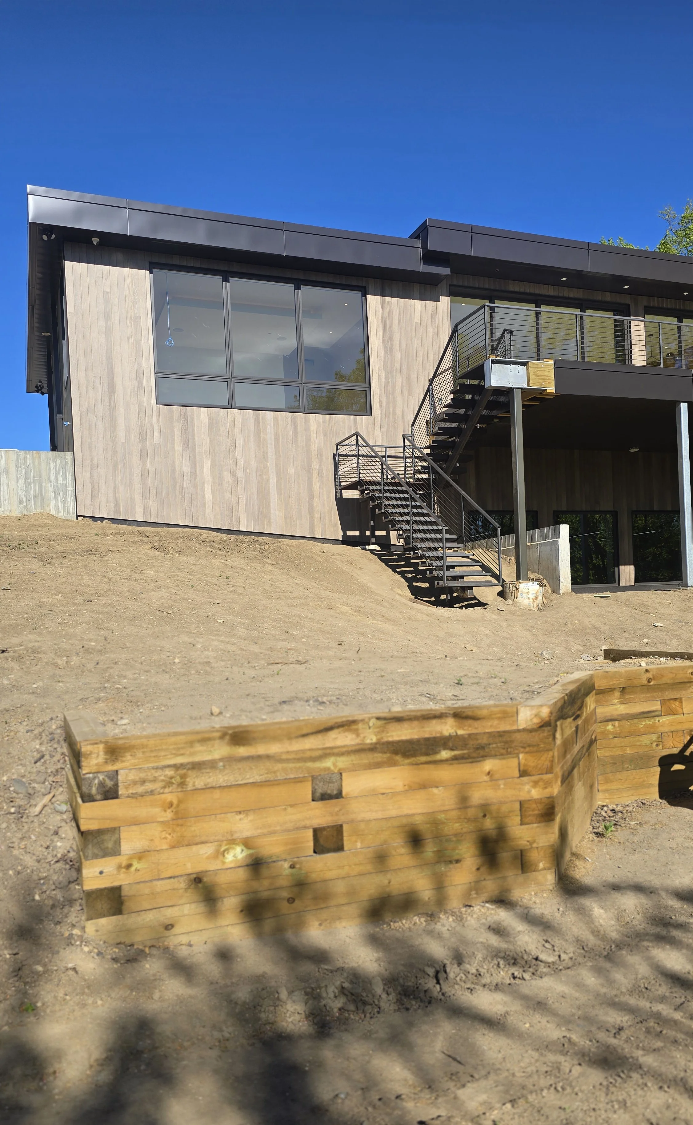 Modern two-story house with large windows, exterior staircase, and balcony, situated on a dirt lot under a clear blue sky.