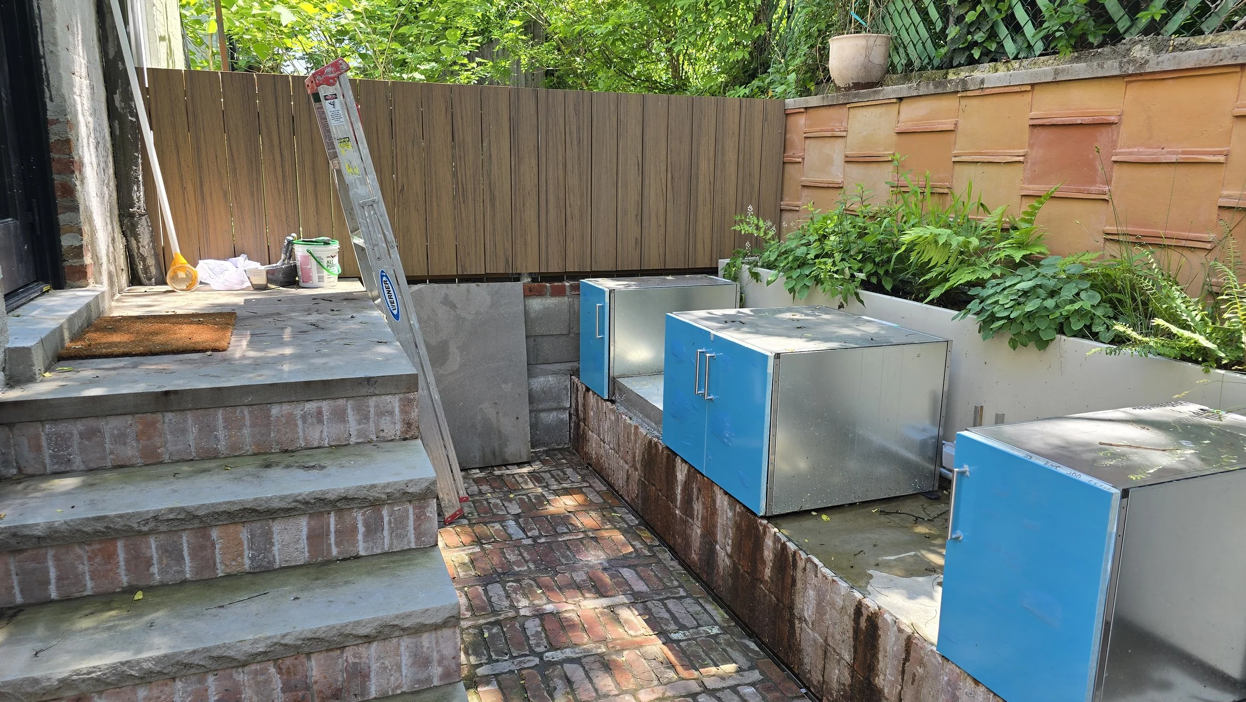 Backyard patio under renovation with brick stairs, a wooden fence, a green garden bed, a ladder, paint cans, and construction materials.