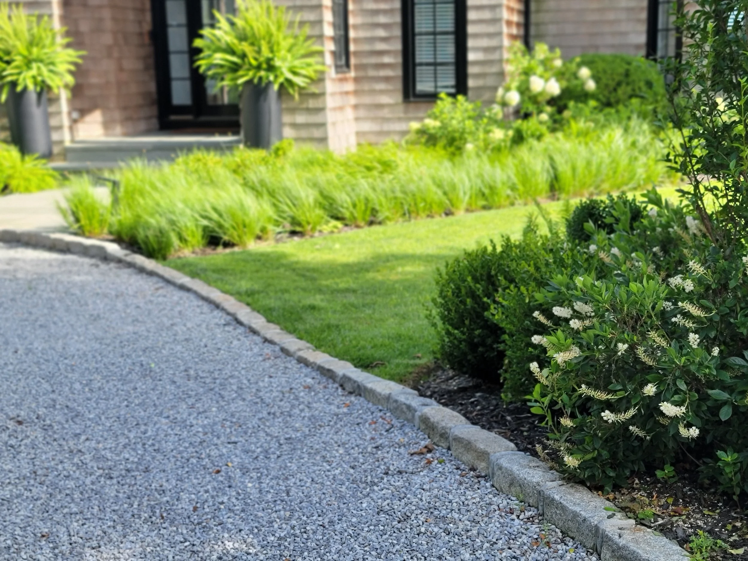 A gravel driveway bordered by a stone edge, with lush green shrubs and plants along the side. The background shows a shingled house with a front door and a window, and some decorative potted plants near the entrance.