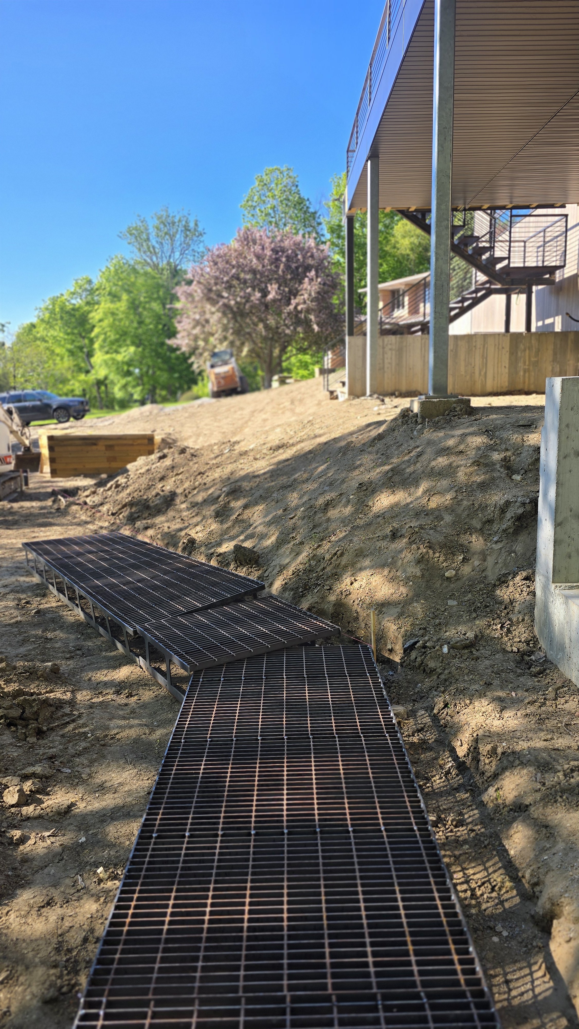 Construction site with metal grates forming a pathway, building framing with steel support columns, dirt ground, trees in background, and a blue sky.
